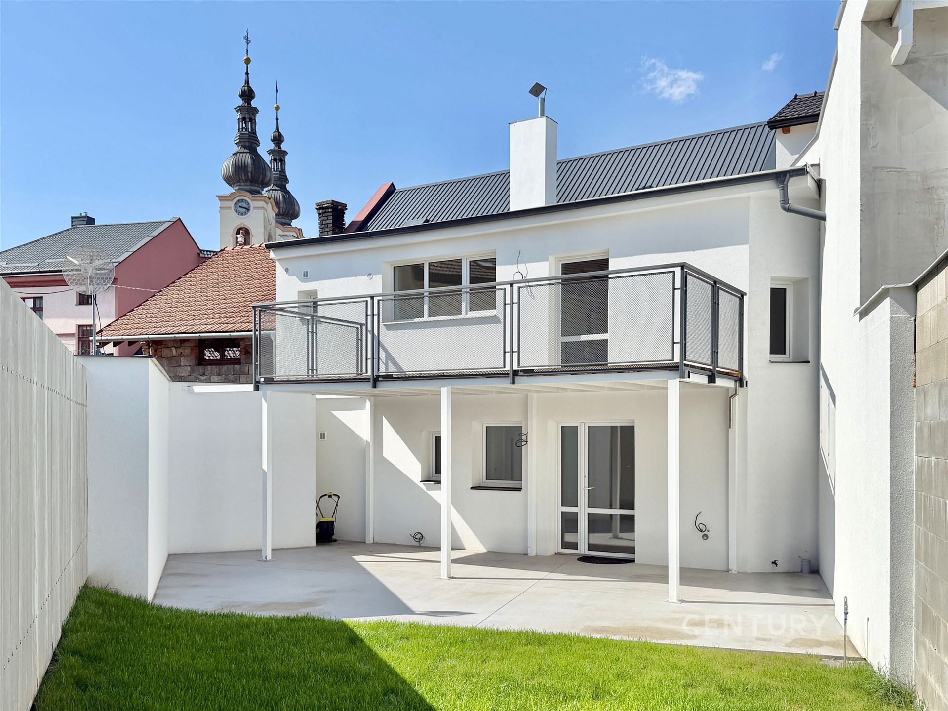 Přízemí domu s moderní fasádou, prostornou terasou a upraveným dvorkem. Dům má velká okna a nachází se ve zrekonstruované oblasti blízko kostela.