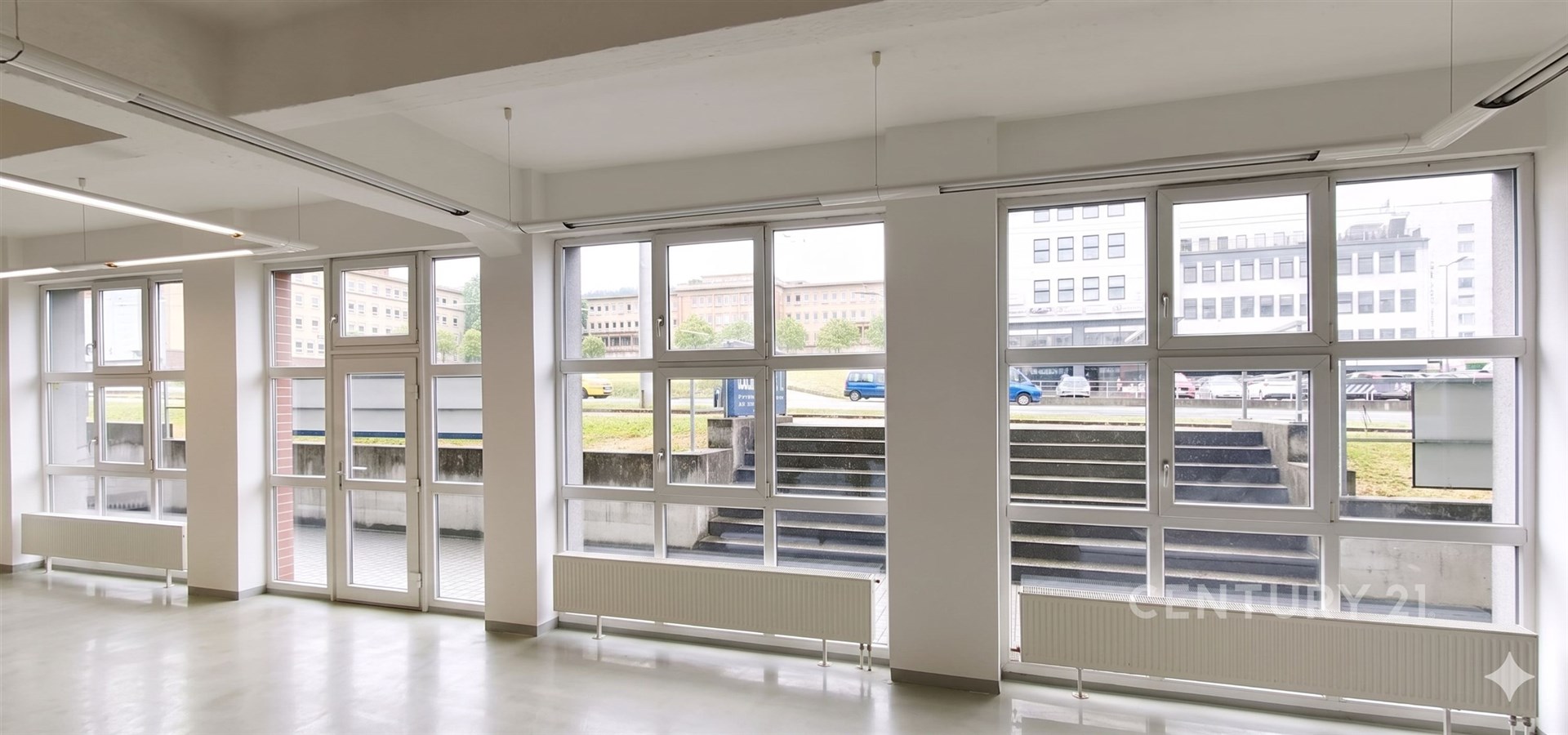 Světlá kancelářská místnost s velkými okny, moderní dlažbou a bílými stěnami, ideální pro kreativní pracovní prostředí.