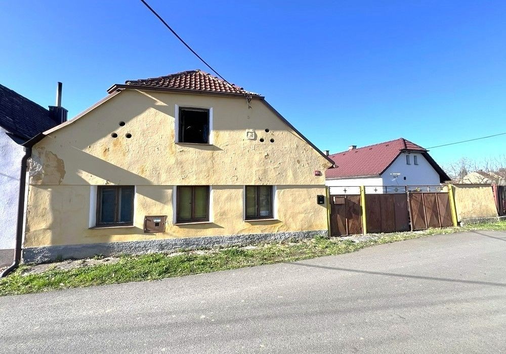 Exteriér venkovského domu s dřevěnými okenicemi a sedlovou střechou, vhodný pro rekonstrukci, nachází se v klidné lokalitě s výhledem na ulici.