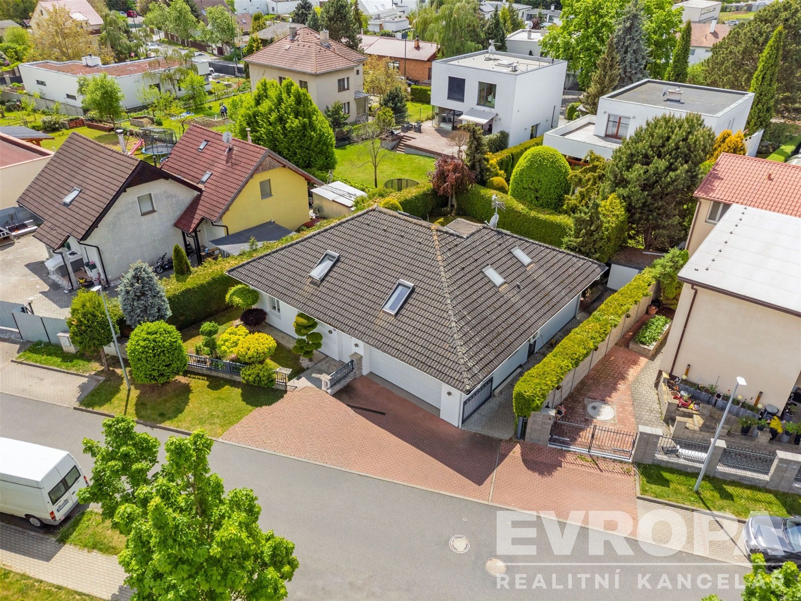 Pohled na moderní bungalov s šikmou střechou a střešními okny, obklopený udržovanou zahradou. Nemovitost se nachází v klidné obytné čtvrti.