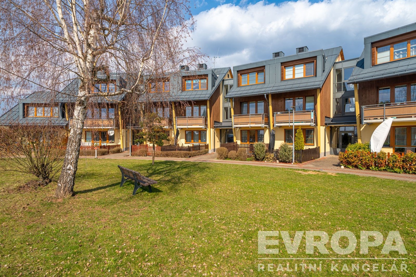 Moderní řadové domy s dřevěnými fasádami a velkými okny, obklopené zeleným trávníkem, nabízejí klidné bydlení v přírodní lokalitě.