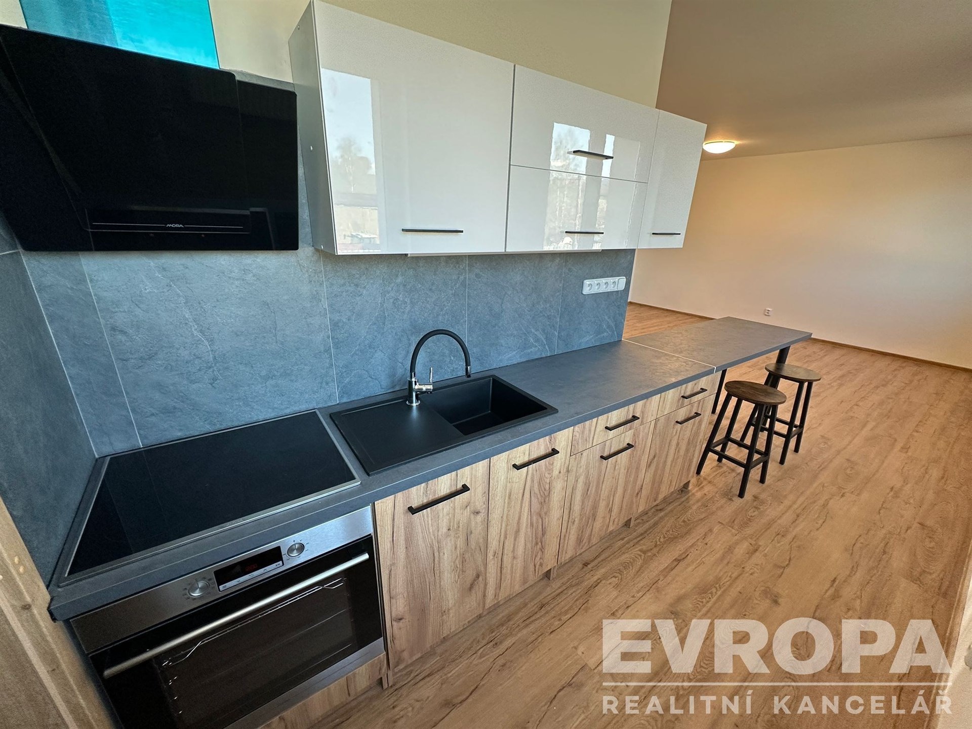 Moderní kuchyň s parketovou podlahou, dřevěnými skříňkami a černými spotřebiči, vedle prostoru pro jídelní stůl či barový pult