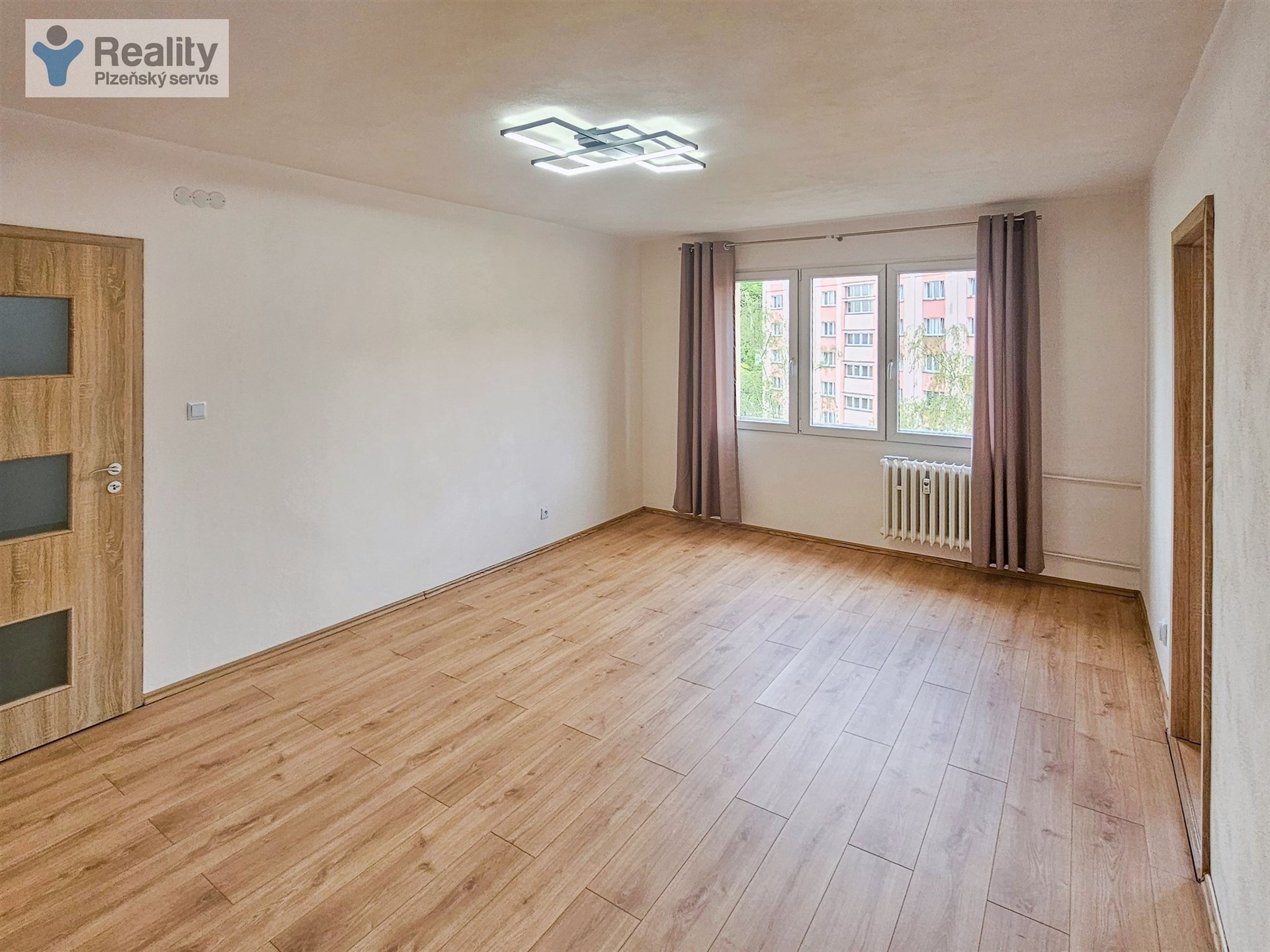 Světlý obývací pokoj s moderními parketovými podlahami, velkými okny a dřevěnými dveřmi, ideální pro rodinné bydlení.