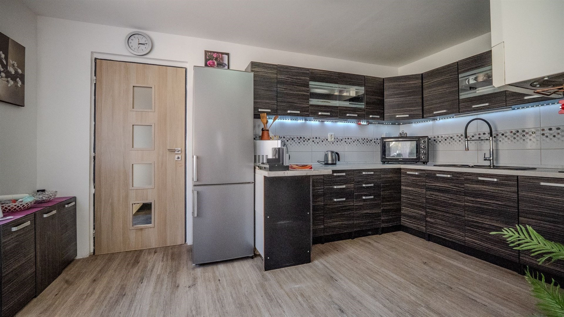 Moderní kuchyň s tmavou dřevěnou linkou, parketovou podlahou a vestavěnými spotřebiči, nabízí světlý a elegantní prostor.