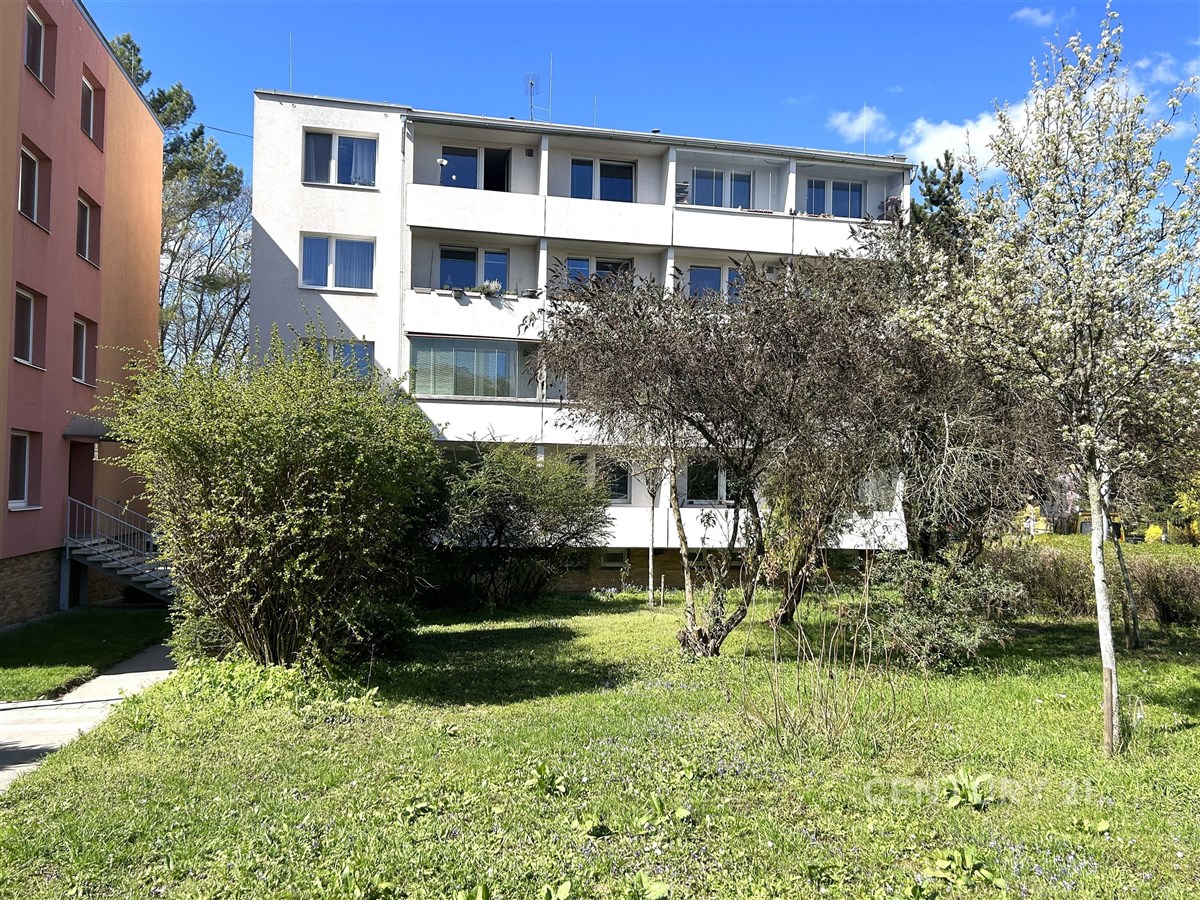 Čtyřpodlažní bytový dům s bílou fasádou, obklopený zeleným trávníkem a stromy, umístěný na klidném místě v rezidenční čtvrti.