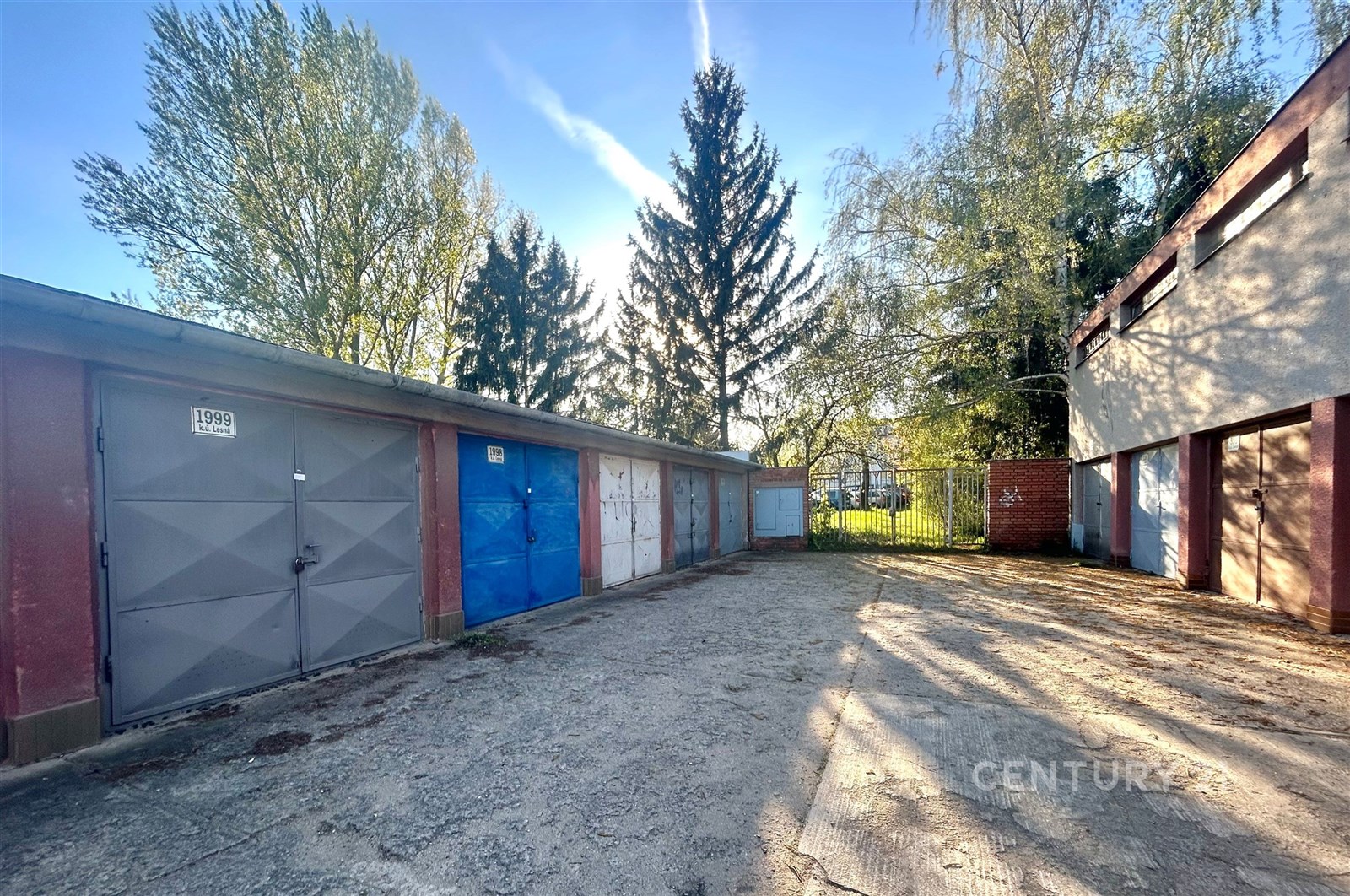 Uzavřená garážová stání s plechovými vraty, klidná lokalita, pevná betonová plocha před garážemi, bezpečný přístup, ideální pro parkování.