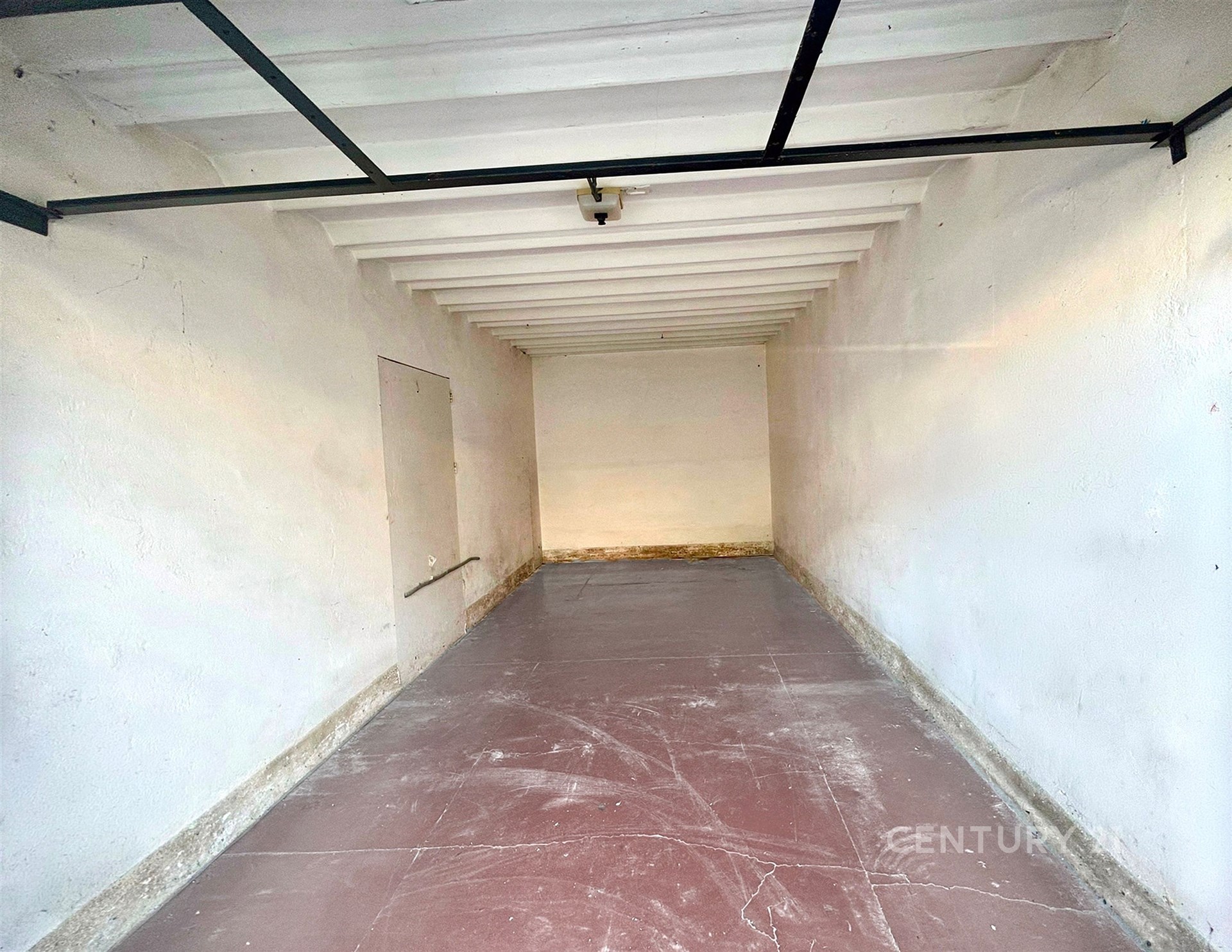Prázdná garáž s betonovou podlahou a bílými stěnami, ideální pro skladování nebo parkování, v původním stavu, bezpečnostní dveře.
