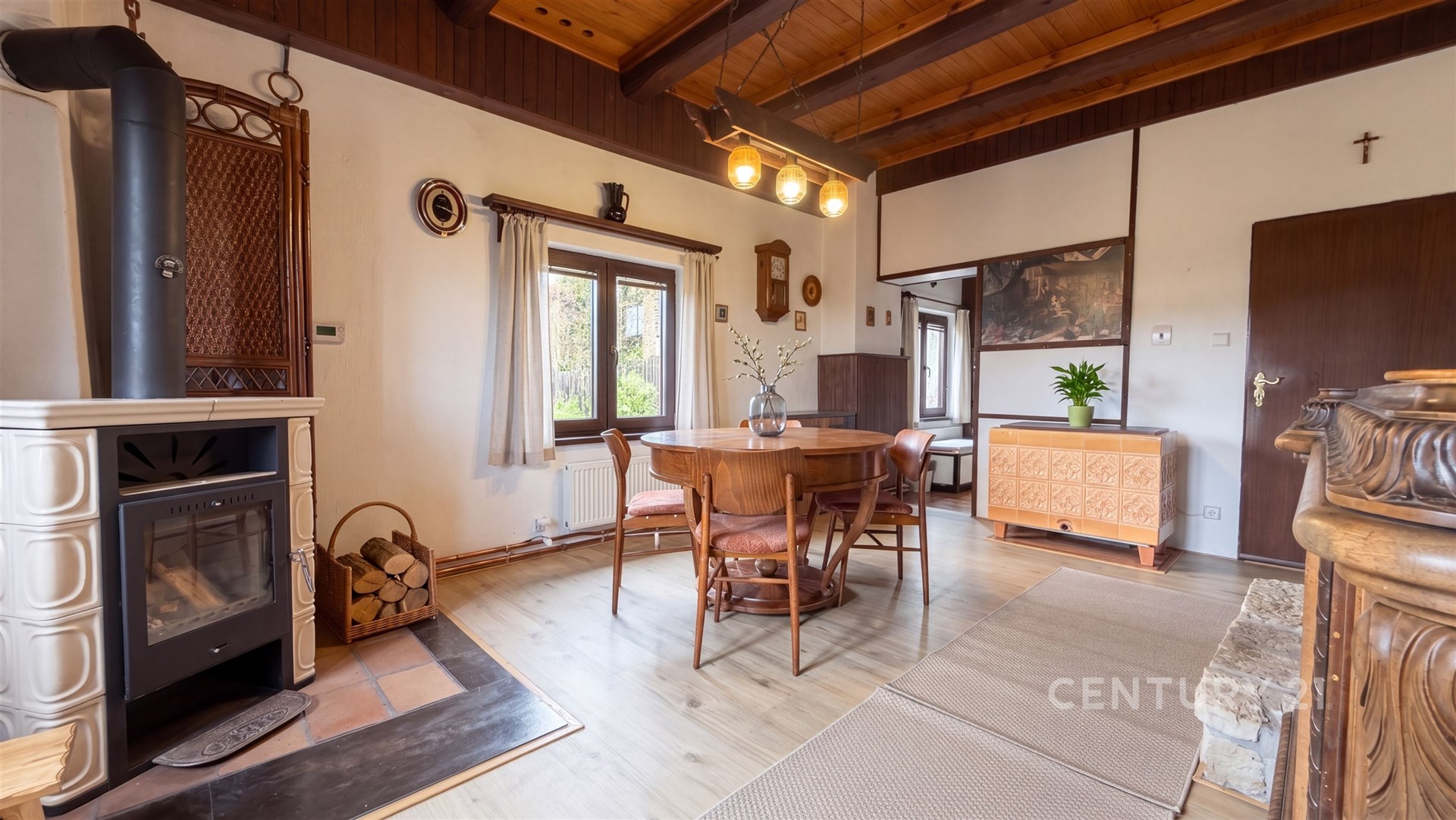 Jídelna s dřevěnými trámy, krbovými kamny a parketovou podlahou nabízí útulný a rustikální styl, doplněný velkými okny pro přirozené světlo.