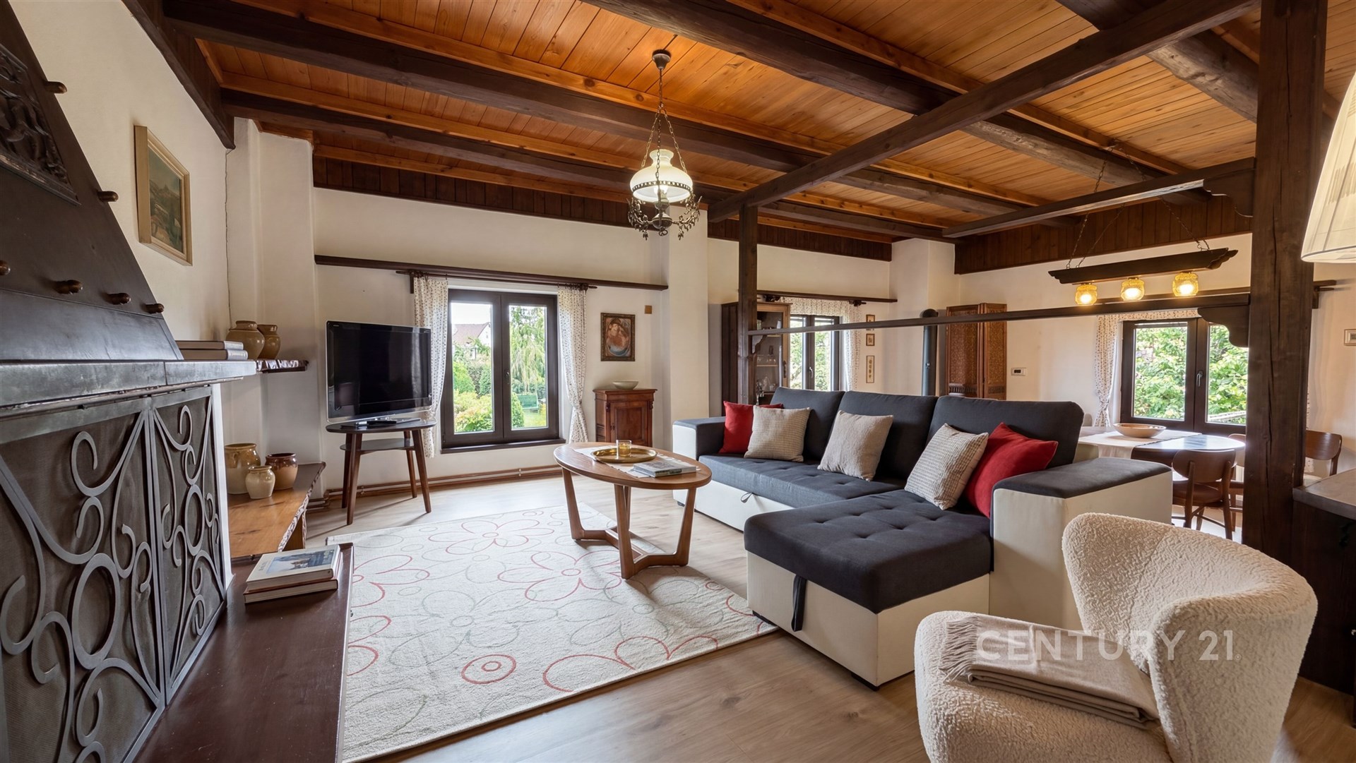 Prostorný obývací pokoj s dřevěnými stropy, parketovou podlahou a velkými okny nabízejícími výhled do zahrady, zařízený v rustikálním stylu.