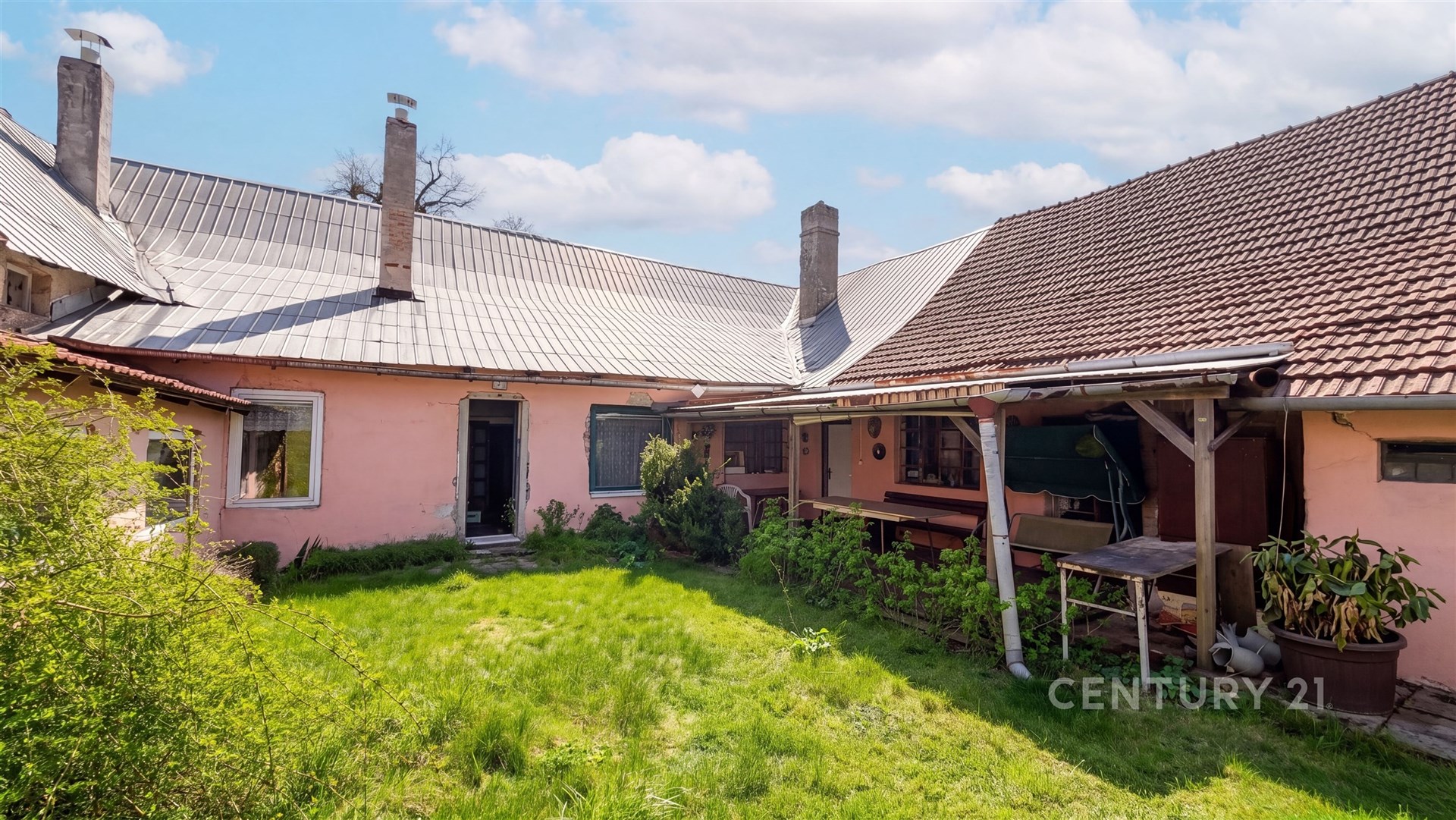 Zahrada s travnatým pozemkem u rodinného domu, s terasou a rozšířeným křídlem, vhodná pro rekonstrukci. Dominantní jsou tradiční střešní krytiny.