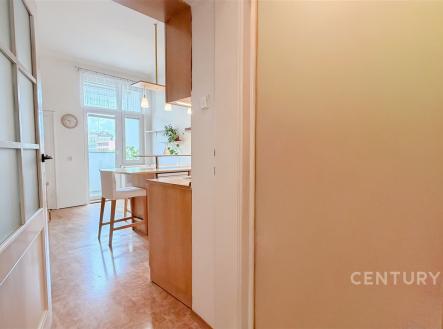 Světlá kuchyň s parketovou podlahou, moderními dřevěnými skříňkami a velkými okny, poskytujícími dostatek přirozeného světla.