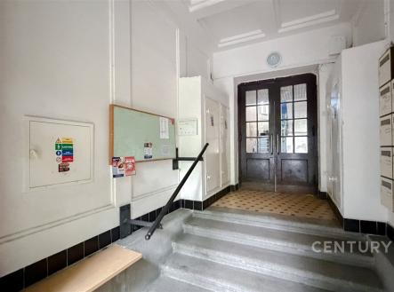 Vstupní hala s dlažbou, dřevěnými dveřmi a poštovními schránkami, ve zrekonstruované budově se světlými stěnami.