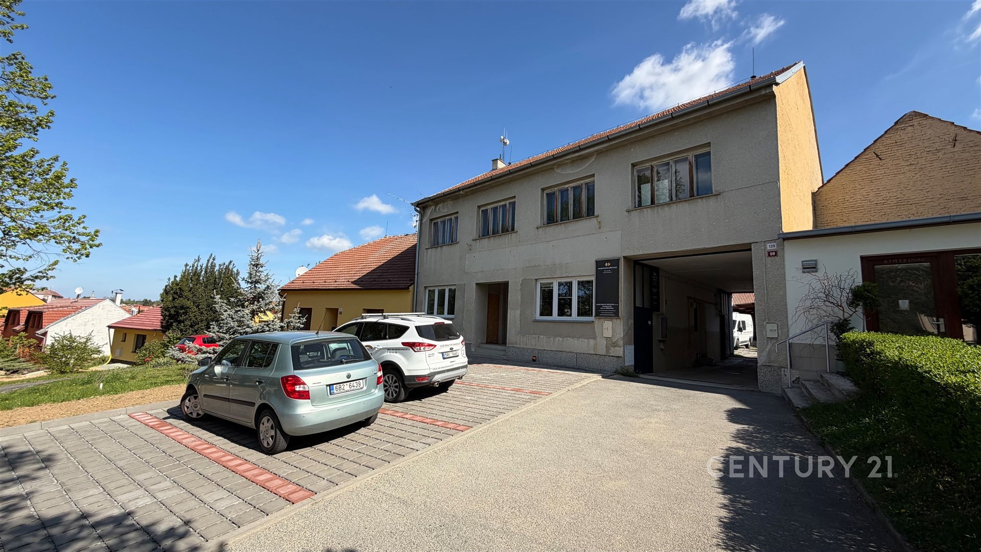 Na dvoře rodinného domu se nachází parkovací prostor s dlažbou a zpevněným povrchem, vedlejší zahrada a světlá fasáda budovy.