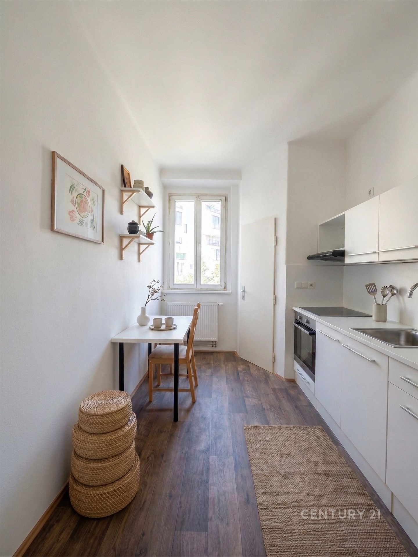 Kompaktní kuchyň s moderními spotřebiči, dřevěnou podlahou a světlými skříňkami, velké okno poskytuje dostatek přirozeného světla.