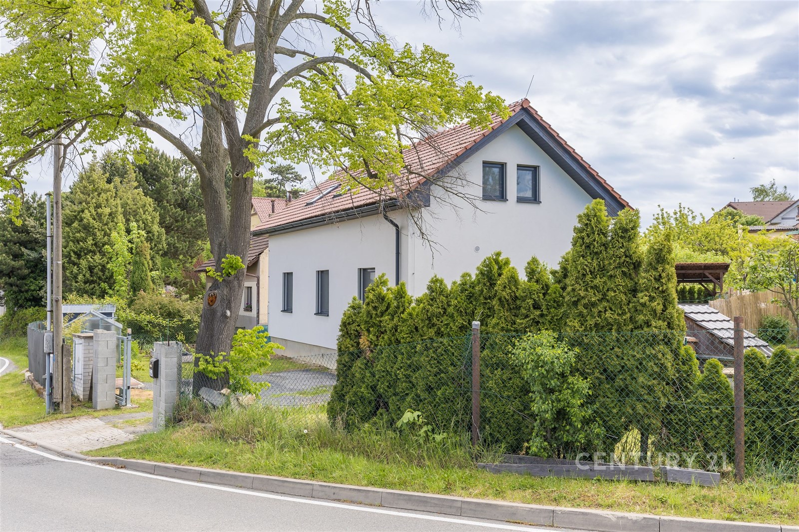 Exteriér rodinného domu s moderní fasádou, šikmou střechou a okny s rámy. Okolní zahrada je udržovaná s živým plotem a zastíněným příjezdem.