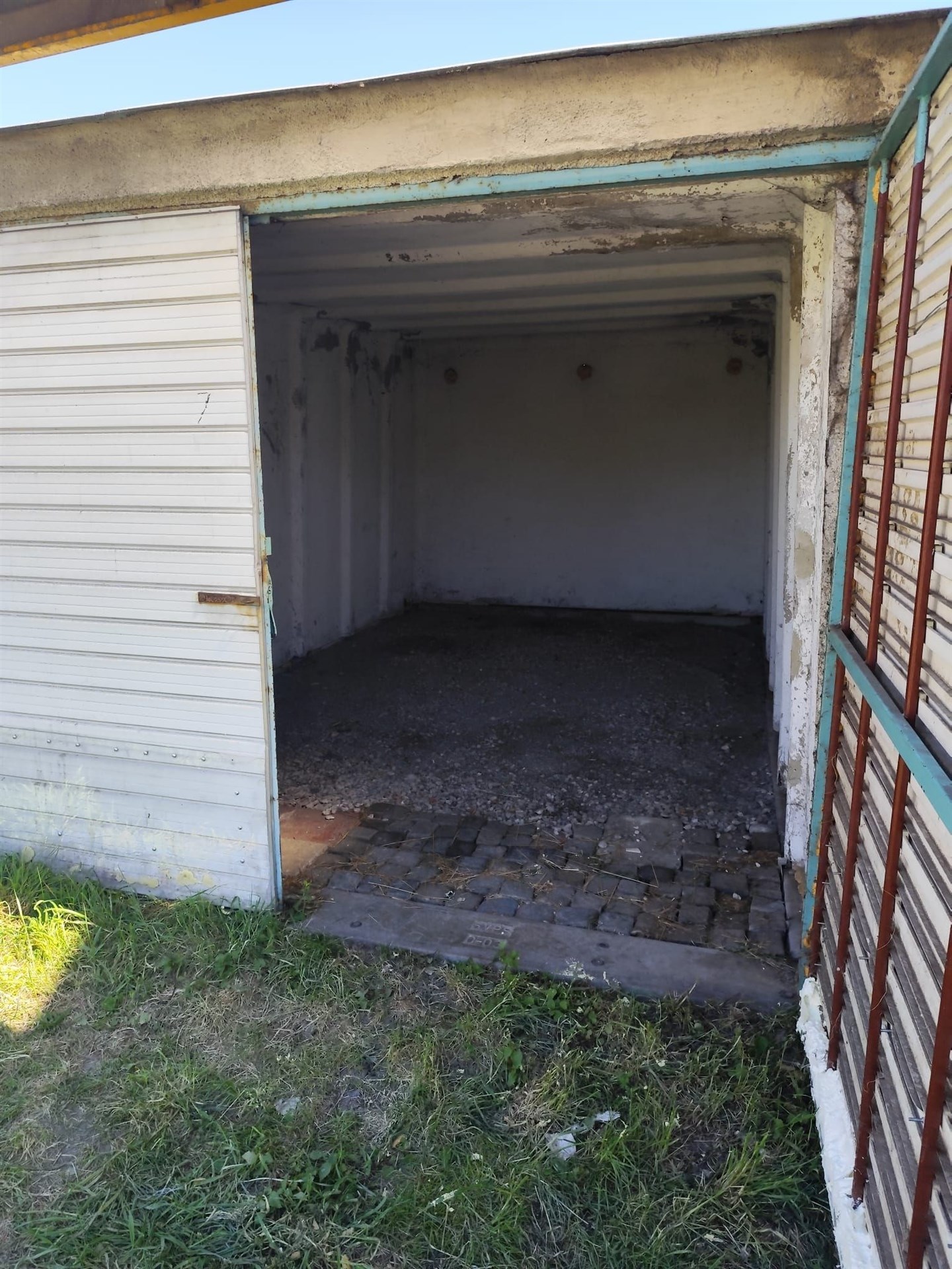 Garážový prostor se štěrkovou podlahou, kovovými vraty a nezrekonstruovaným interiérem.