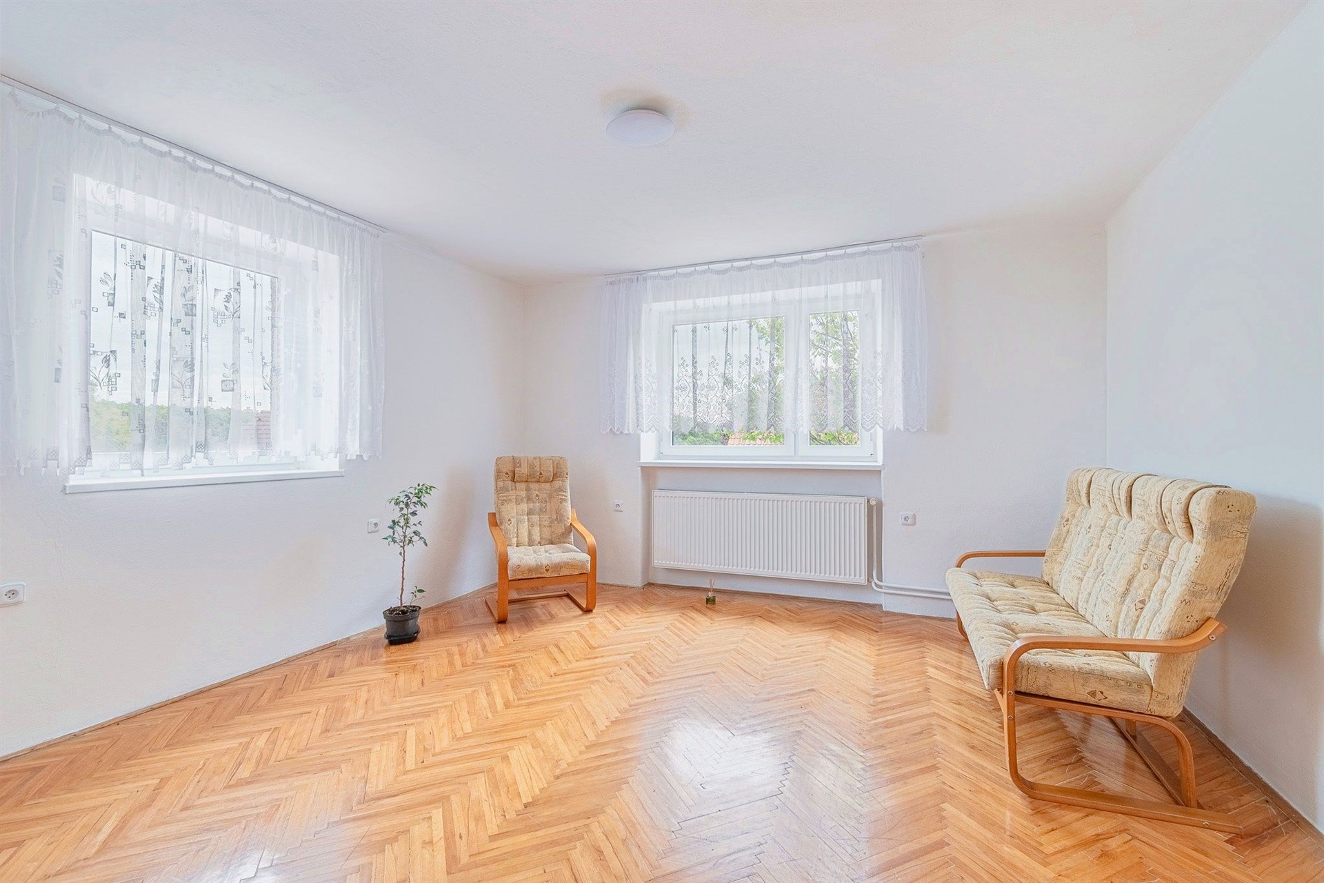 Světlý obývací pokoj s parketovou podlahou, velká okna s bílými závěsy, moderní výmalba a minimalistický nábytek pro pohodlné bydlení.