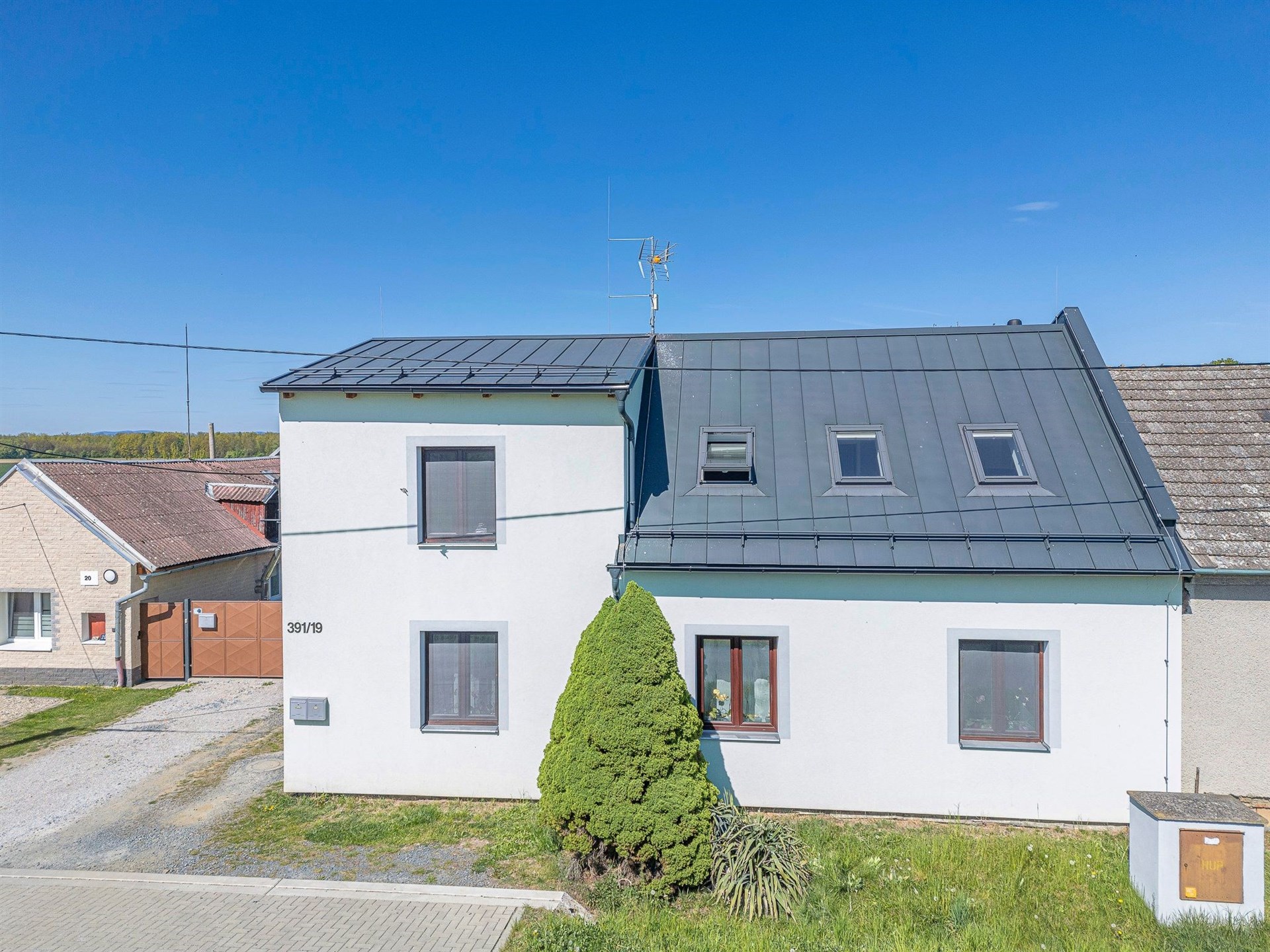 Renovovaná fasáda rodinného domu s moderní střechou, střešními okny a udržovanou zahradou na klidné ulici v malebném venkovském prostředí.
