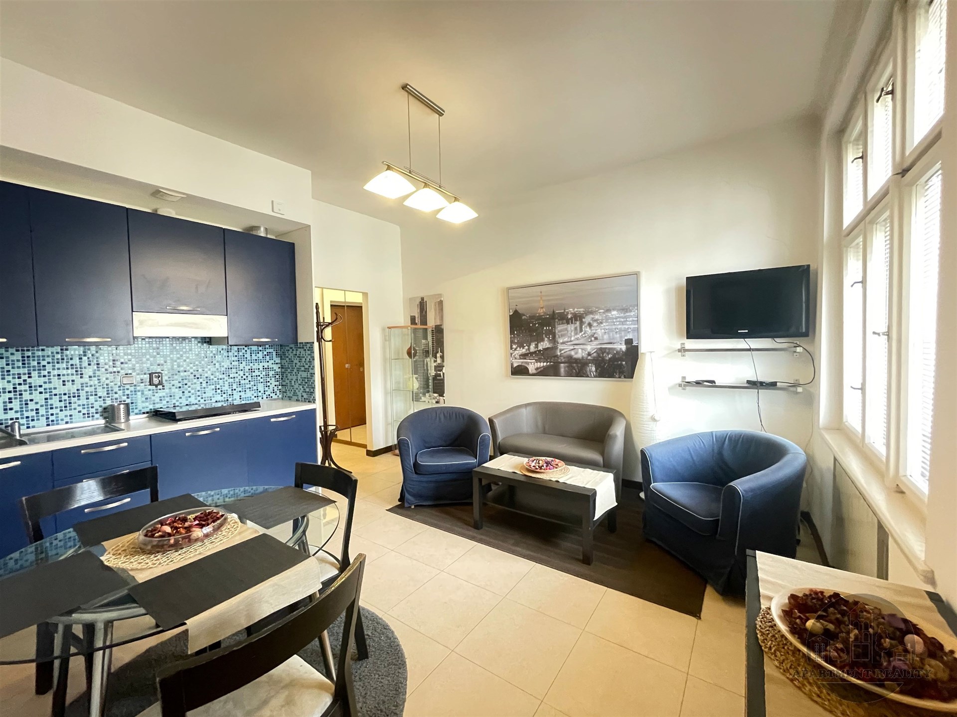 Kuchyňský kout s tmavě modrými skříňkami, světlá dlažba a moderní osvětlení, vedle obývací zóny s pohovkami a velkými okny.