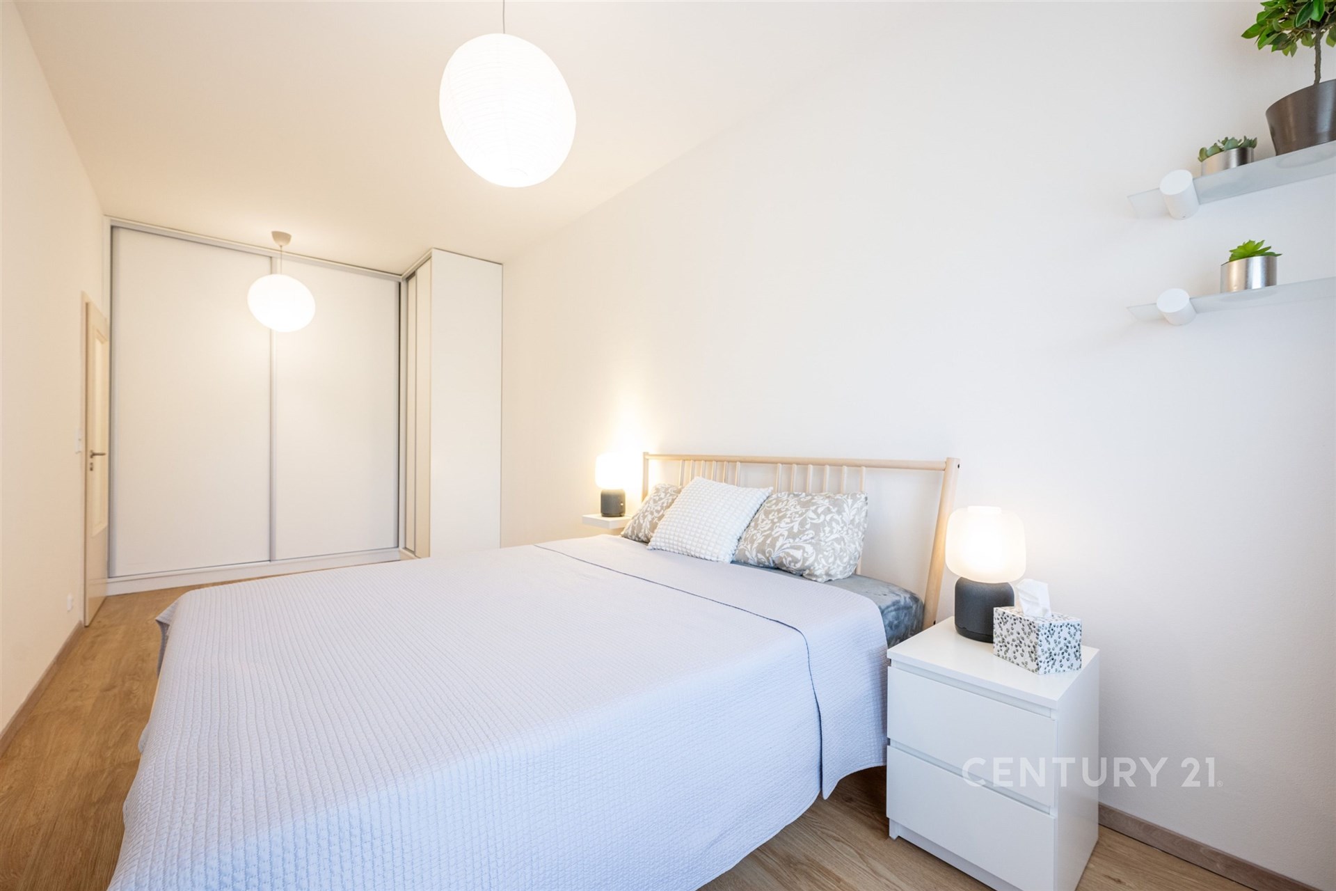 Světlá ložnice s parketovou podlahou, vestavěnými skříněmi a moderním osvětlením. Botanikální dekorace a neutrální tóny zvyšují útulnost.