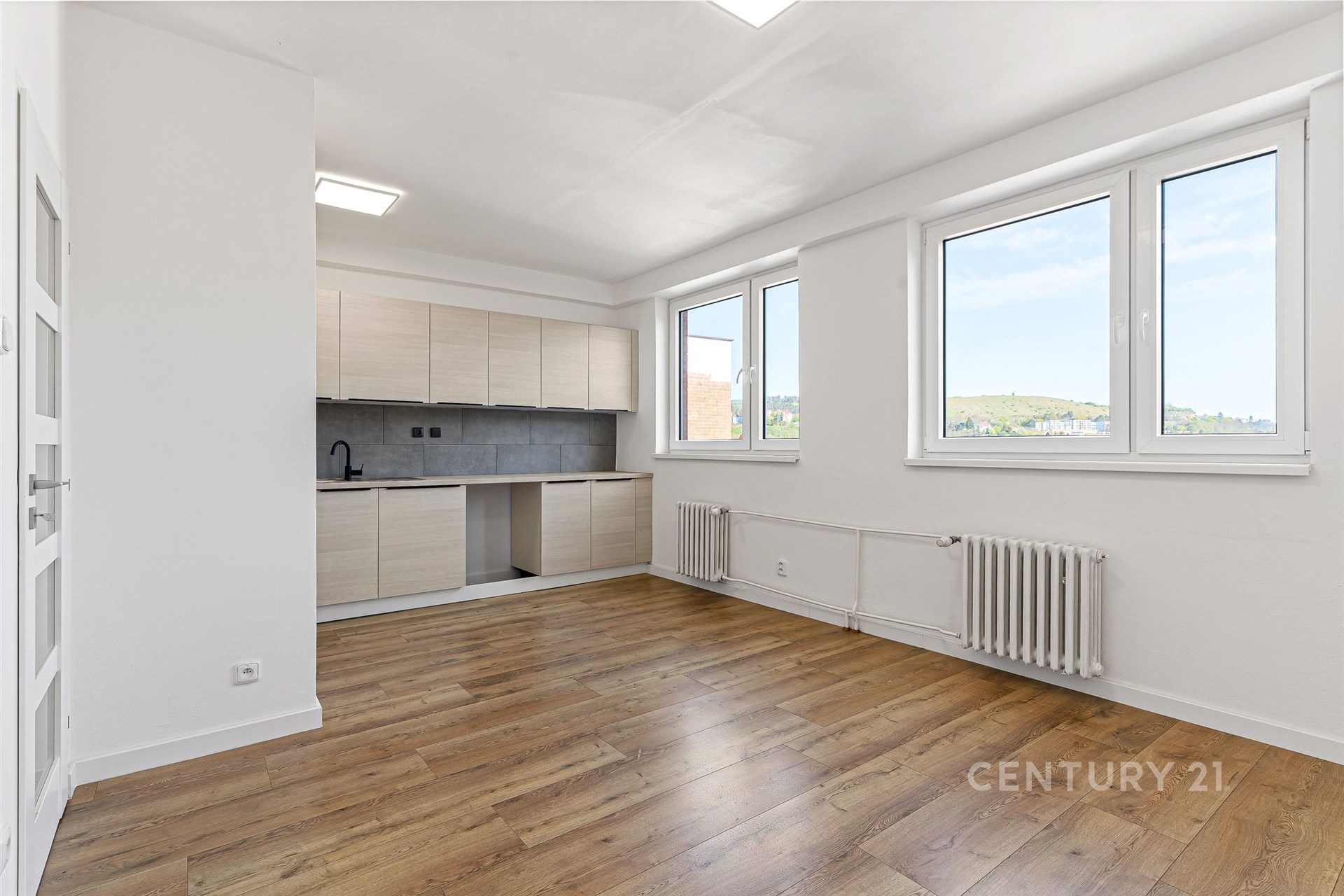 Světlý obývací pokoj s moderní kuchyní, parketovou podlahou a velkými okny. Renovovaný prostor nabízí výhled do krajiny.