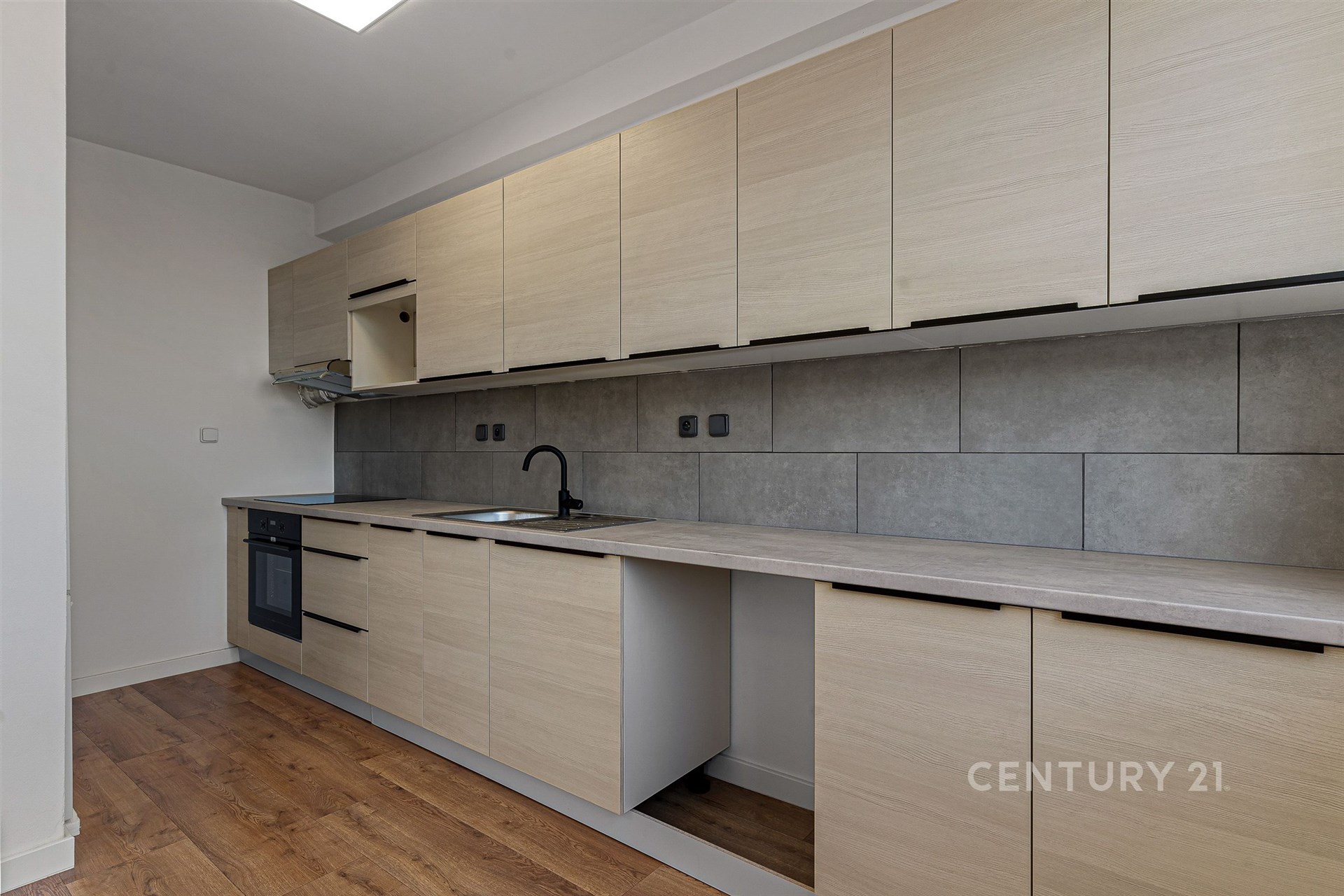 Moderní kuchyň s parketovou podlahou, světlými dřevěnými skříňkami a elektrickou troubou, šedá dlažba na stěně dodává elegantní vzhled.