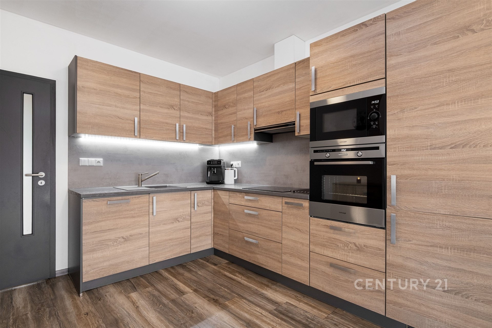 Moderní kuchyň s dřevěnými skříňkami, vestavěnými spotřebiči a parketovou podlahou. Světlý prostor s elegantním designem vhodný pro rodinu.