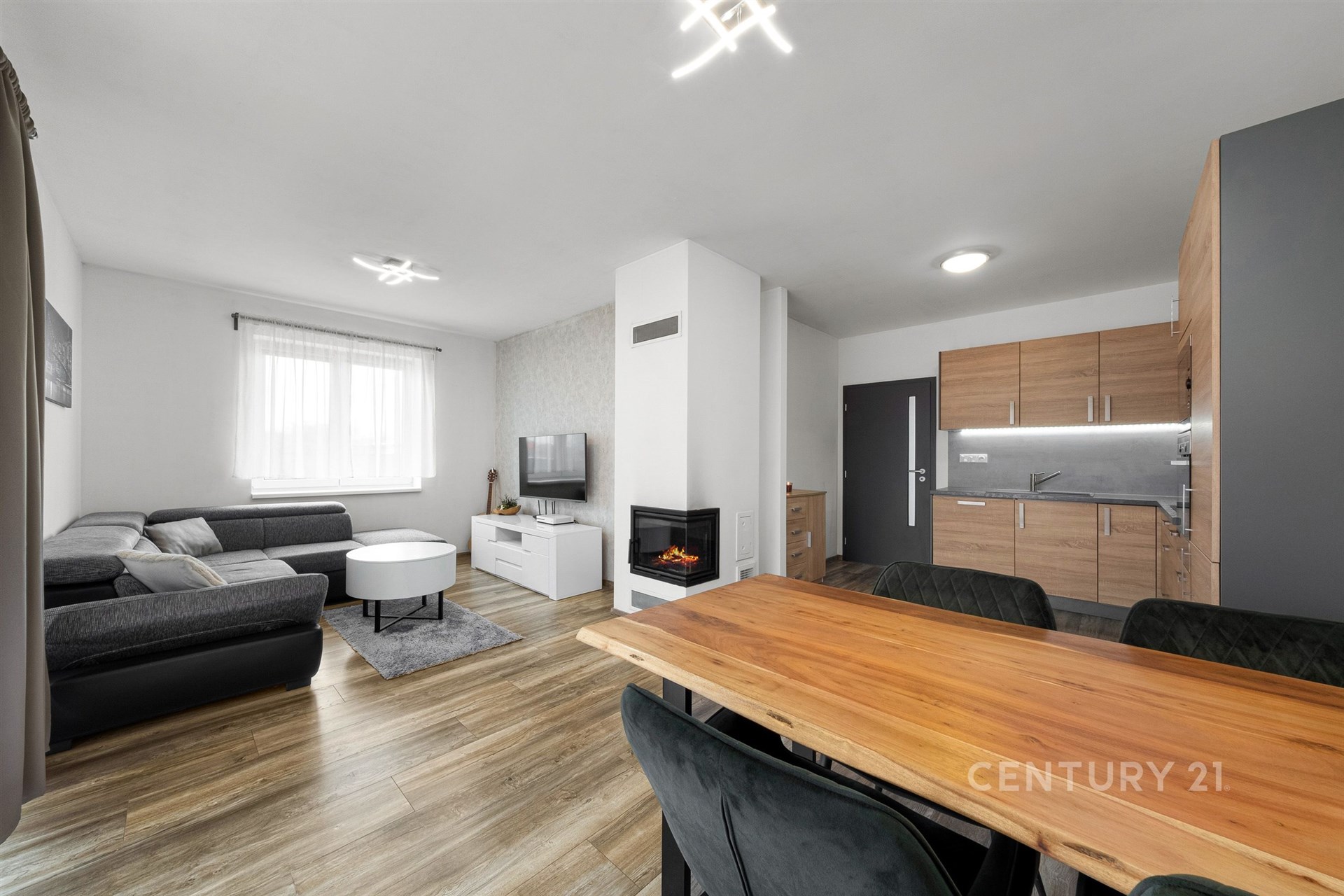 Světlý obývací pokoj s parketovou podlahou, moderní krbová kamna a velká okna, navazuje na kuchyňský kout s dřevěnými skříňkami.