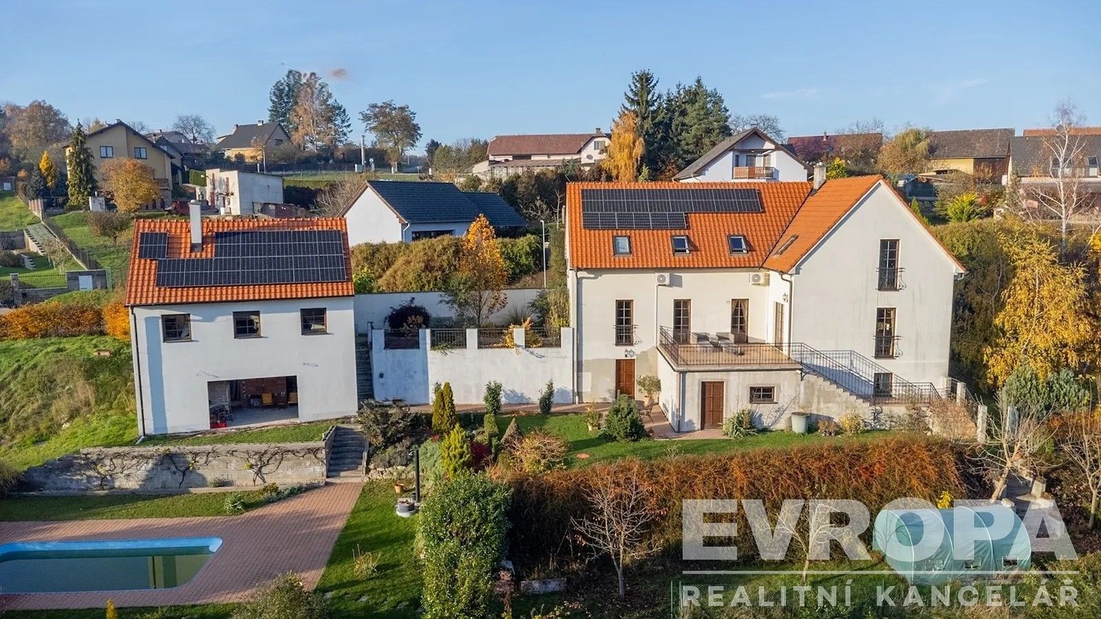 Rodinný dům s červenou střechou, solárními panely a udržovanou zahradou. Moderní architektura s venkovským nádechem a bazénem.