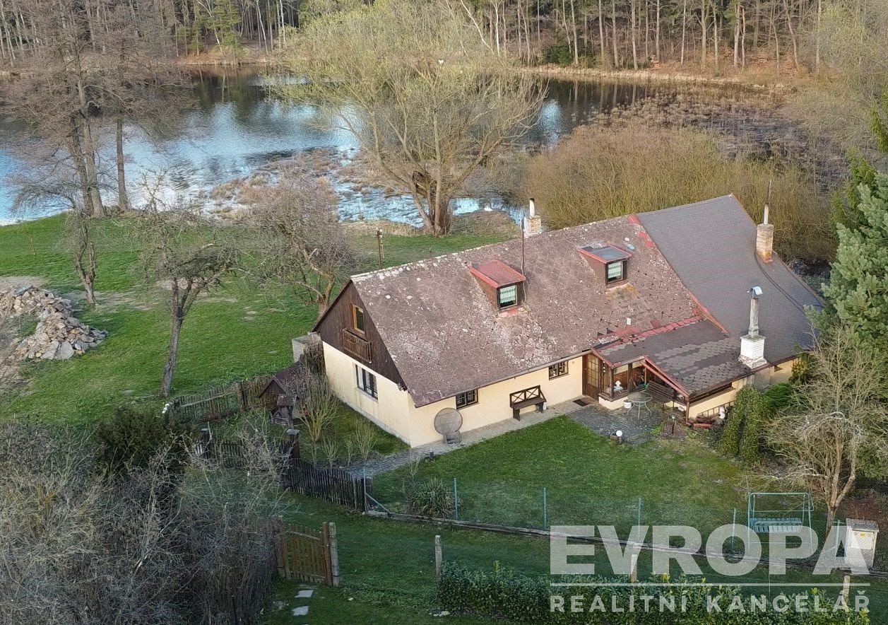 Rustikální venkovský dům u vodní plochy, obklopený zahradou. Klasická střecha, dřevěné obložení, tichá přírodní lokalita.