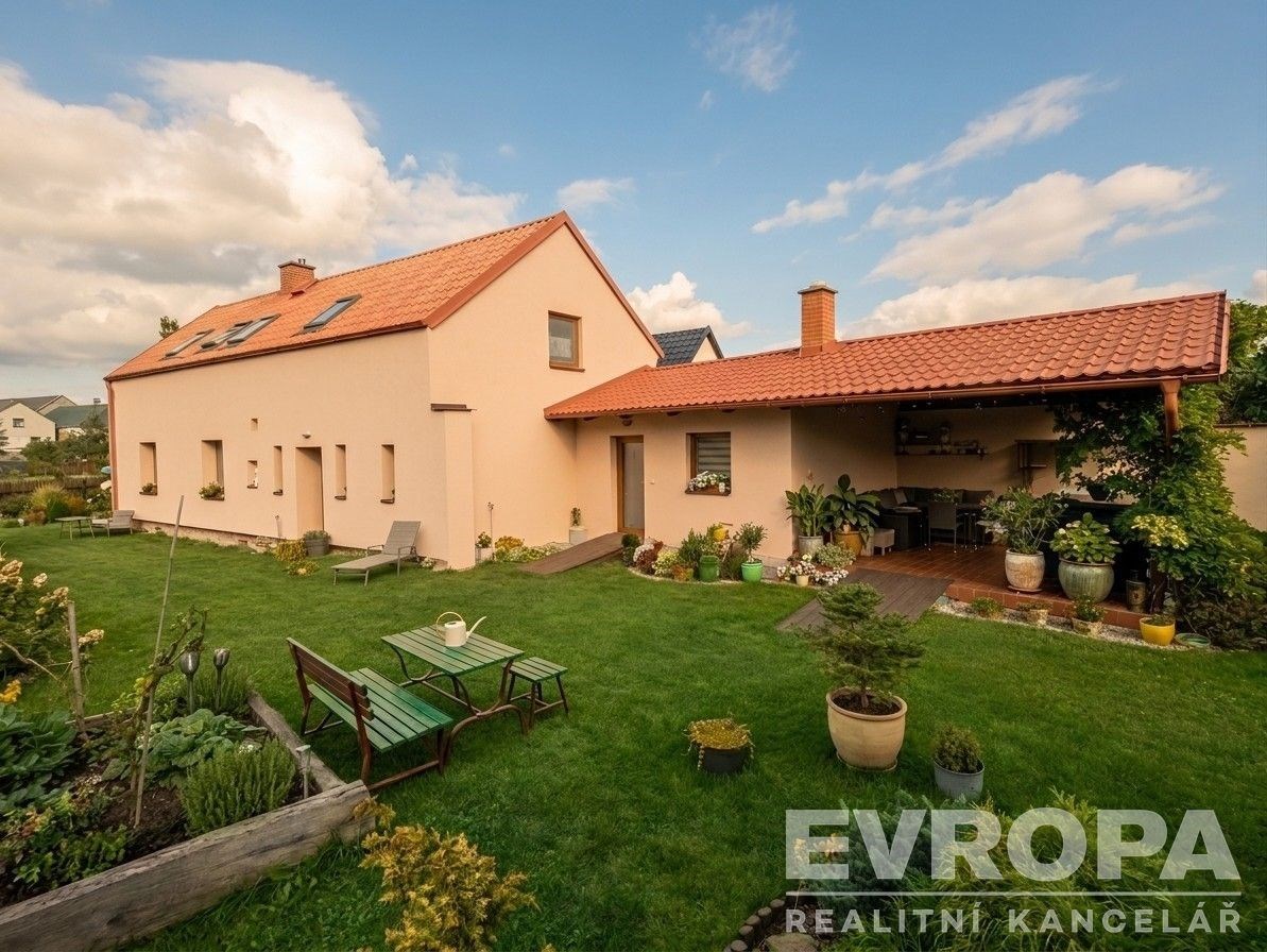 Zahrada u rodinného domu s pečlivě udržovaným trávníkem, zahradní nábytek a pergola. Střecha domu s červenou taškou, klidné okolí.