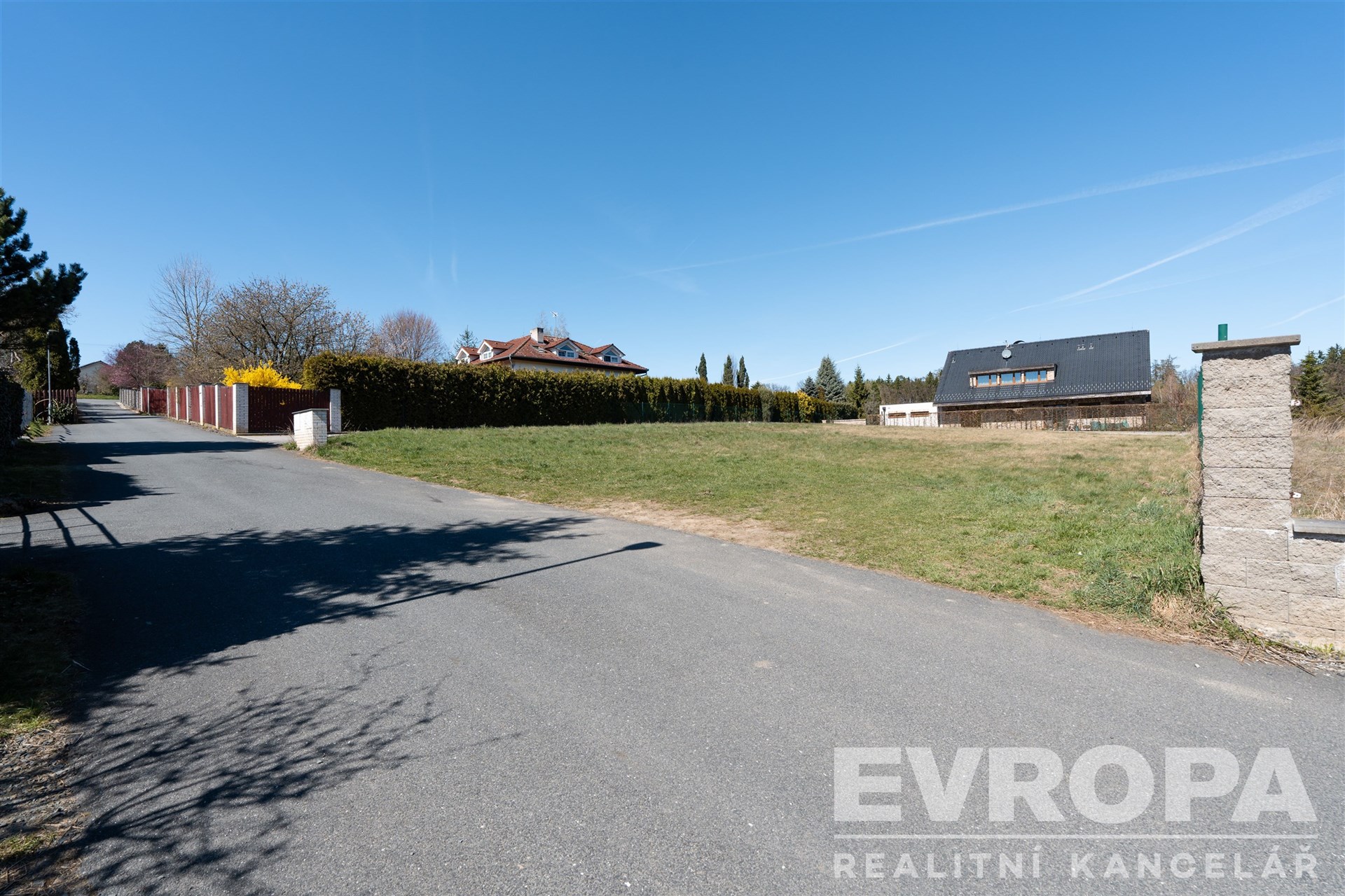 Prázdný pozemek na prodej s asfaltovou příjezdovou cestou, lemovaný živým plotem, ideální pro stavbu domu v klidné lokalitě.