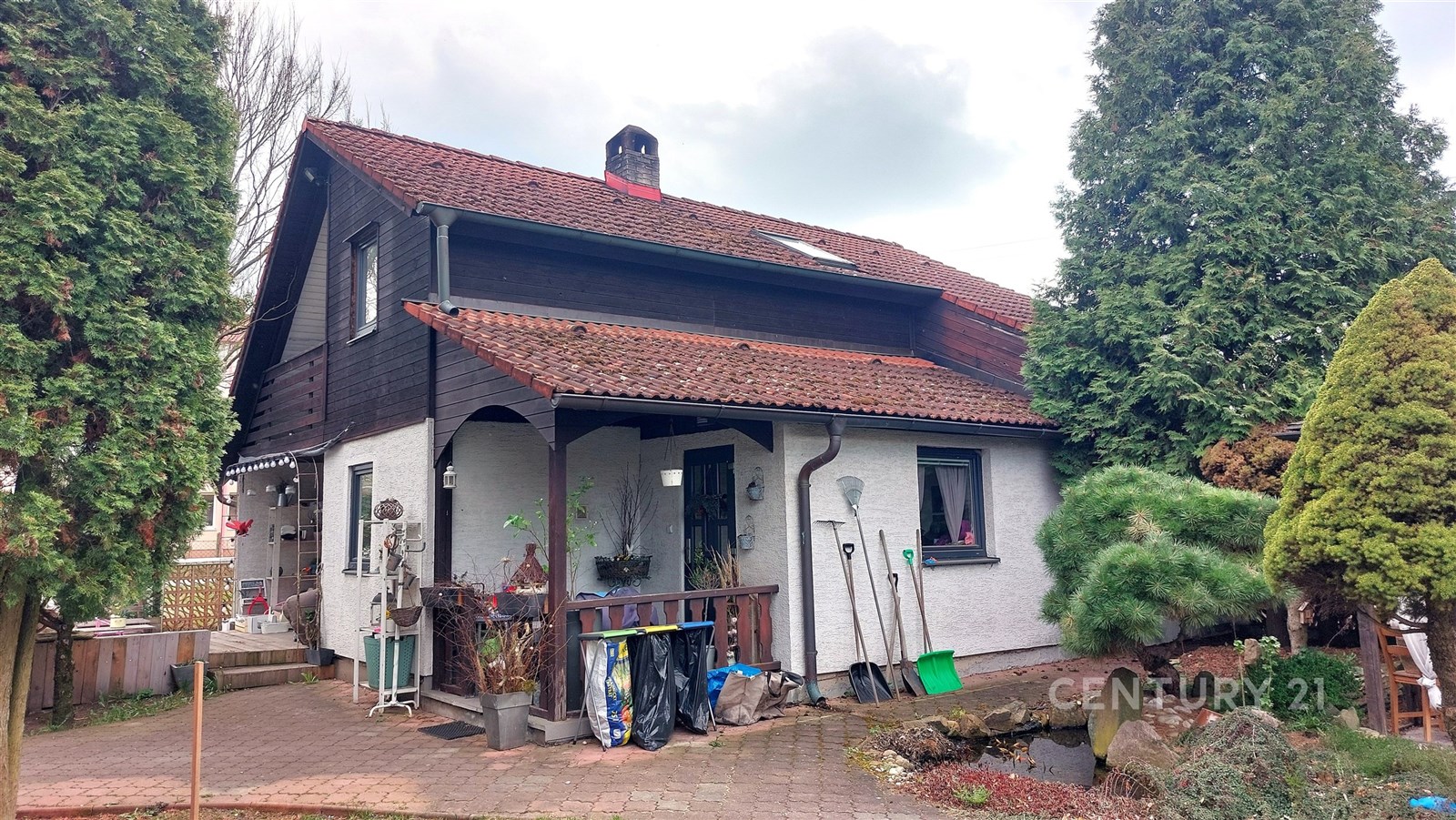 Rodinný dům se šikmou střechou a cihlovými taškami, obložený dřevem. Prostorná zahrada s chodníkem a okrasnými stromy.