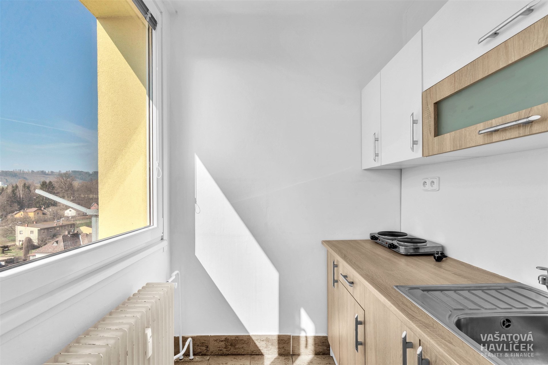 Malá kuchyň s moderními skříňkami, parketovou podlahou a velkým oknem poskytujícím přirozené světlo.