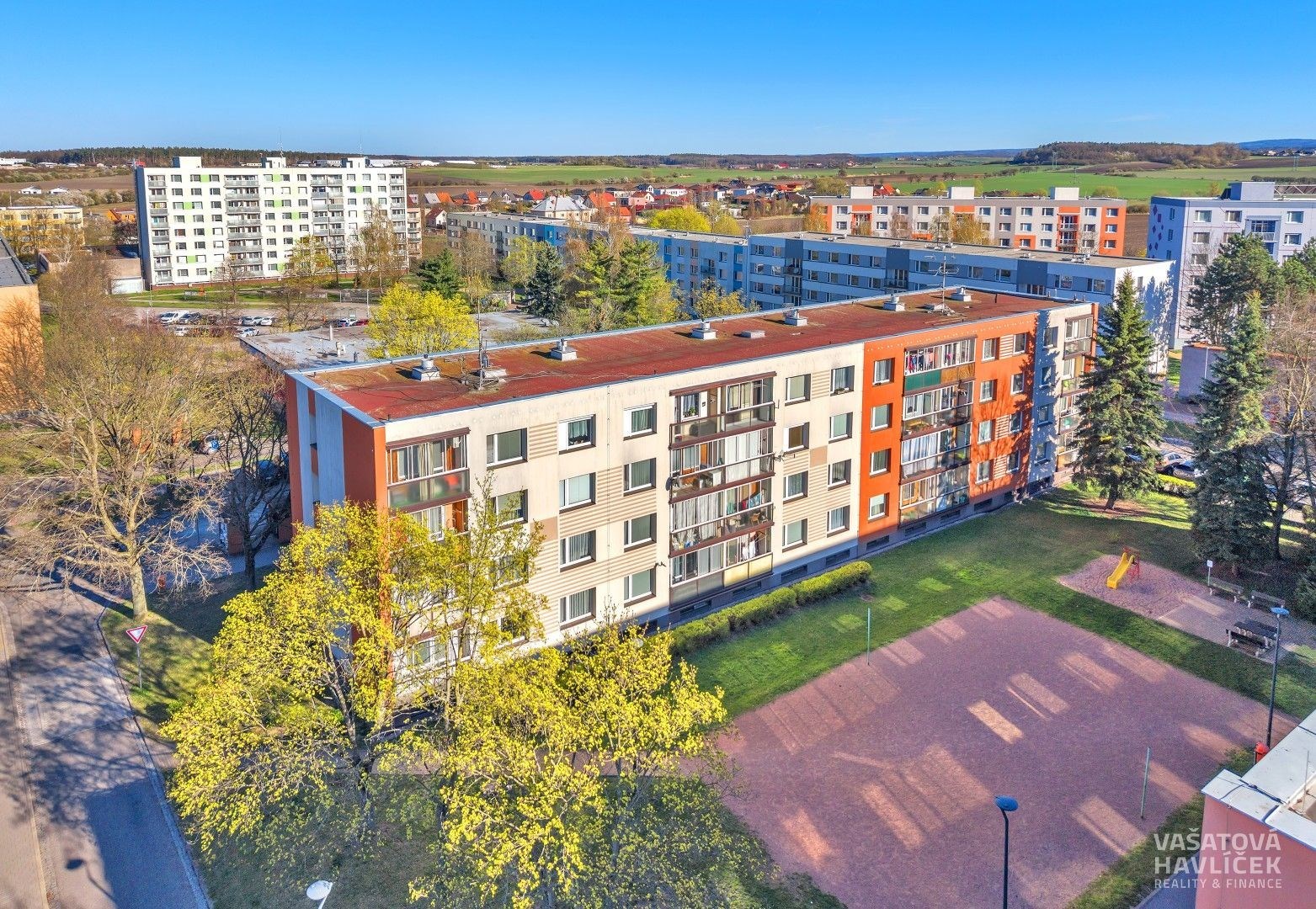 Venkovní pohled na bytový dům s moderní fasádou a novými okny, okolní upravená zeleň a blízké parkoviště poskytují příjemné bydlení.