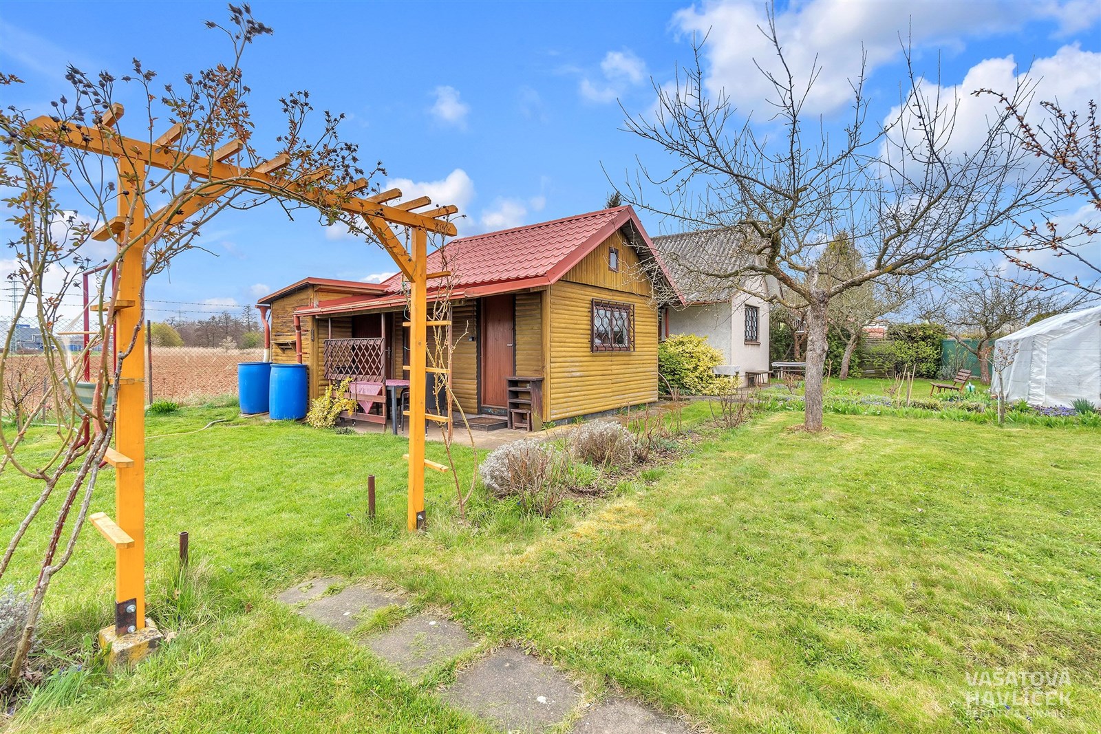 Zahradní domek na rozlehlém pozemku s udržovaným trávníkem, dřevěnými prvky a ovocnými stromy, ideální pro rekreační využití.