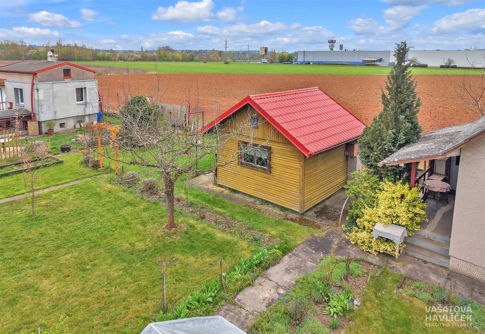 Zahrada s chatkou a terasou, obklopená zelení a záhony. Dřevěná stavba s červenou střechou, vhodná pro rekreaci a relaxaci.