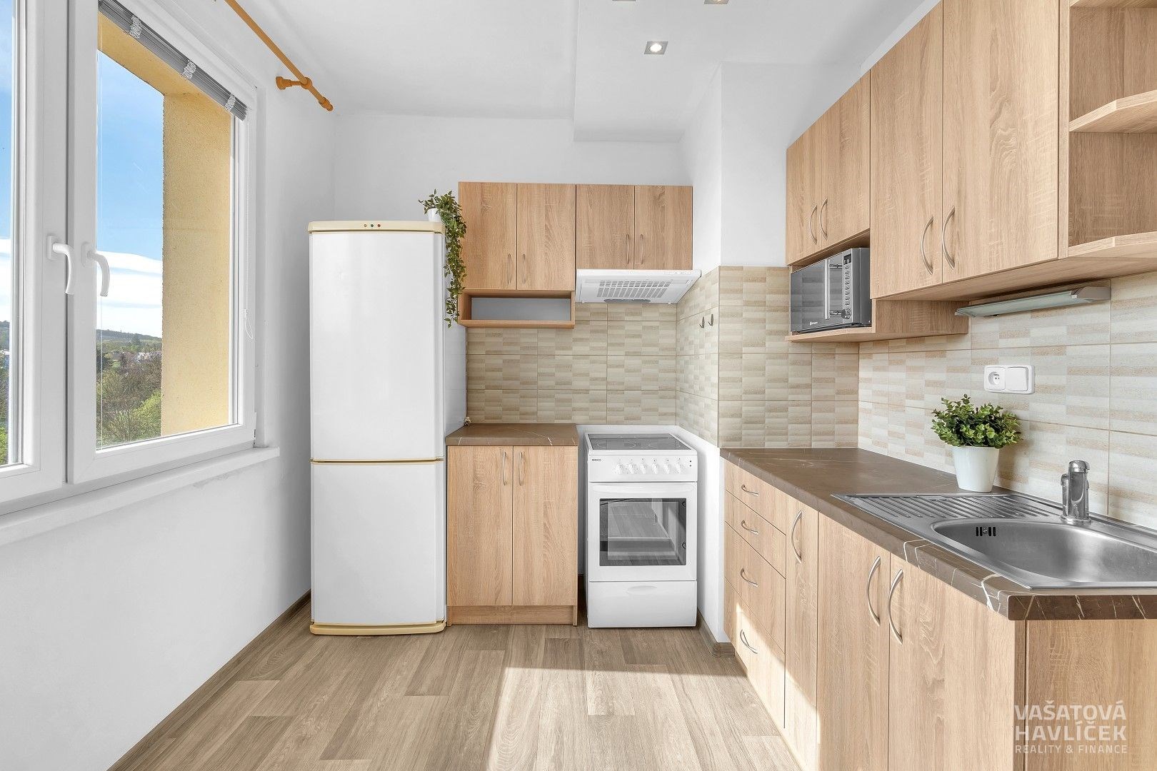 Moderní kuchyň s dřevěnými skříňkami, parketovou podlahou a velkými okny nabízí světlý prostor pro vaření.