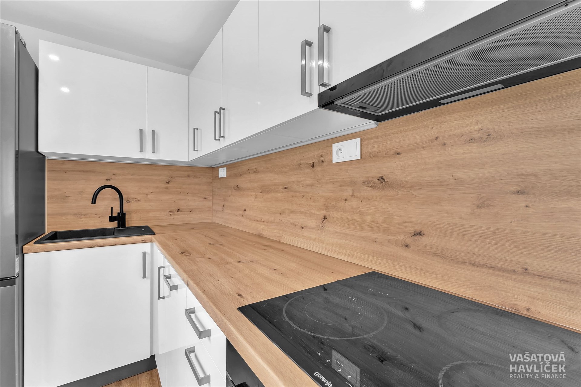 Moderní kuchyň s dřevěnou pracovní deskou, bílými lesklými skříňkami a vestavěnou varnou deskou.