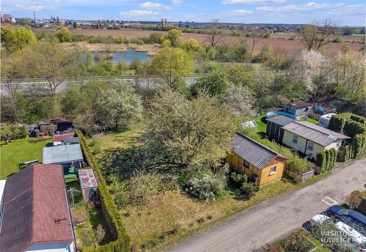 Zahrada s rekreační chatkou, dřevěná konstrukce, klidná lokalita u rybníka, ideální pro odpočinek, obklopená vzrostlými stromy a zelení.