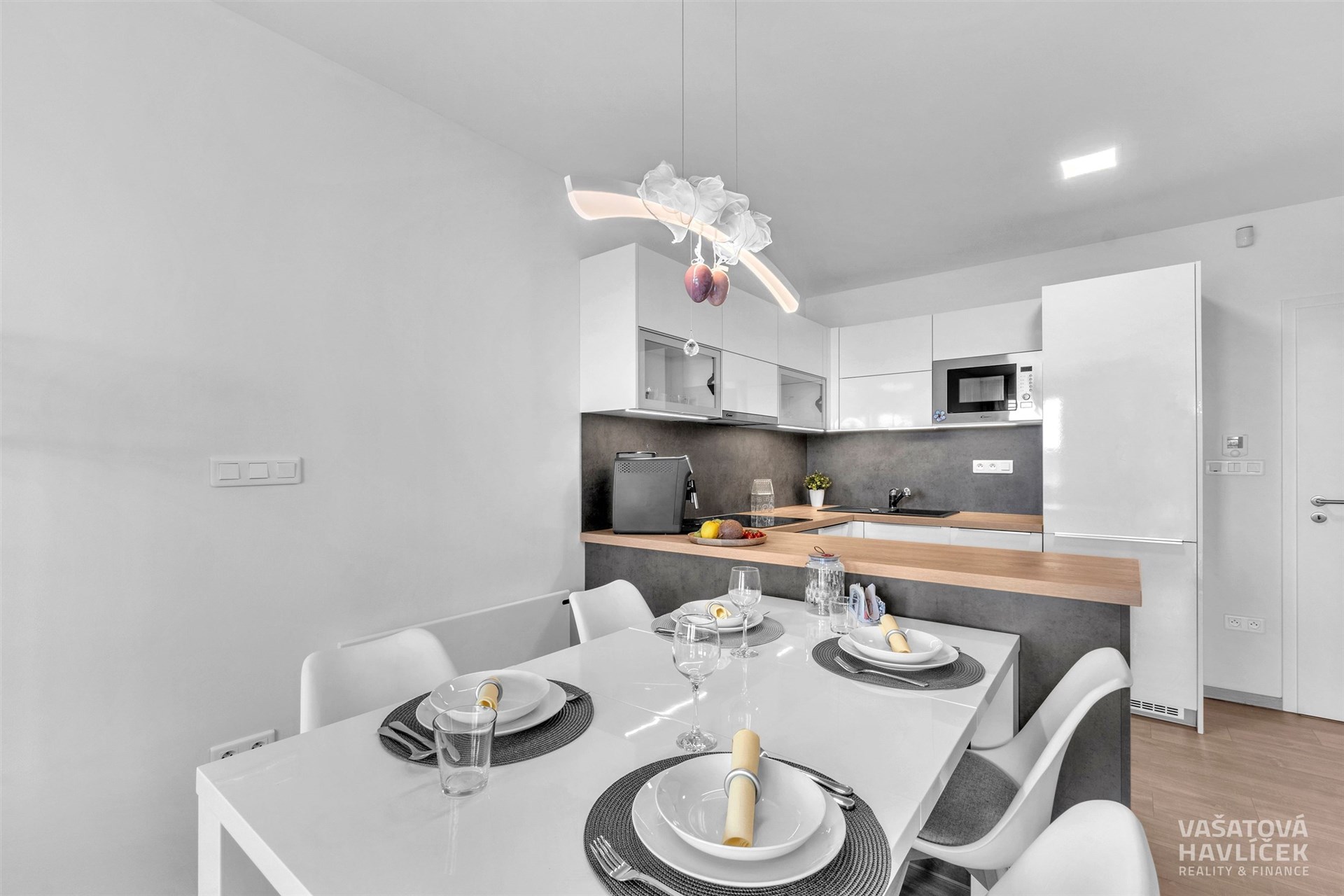 Moderní kuchyň s vestavěnými spotřebiči, světlou pracovní deskou a jídelním stolem. Bílé skříňky a minimalistický design poskytují elegantní vzhled.