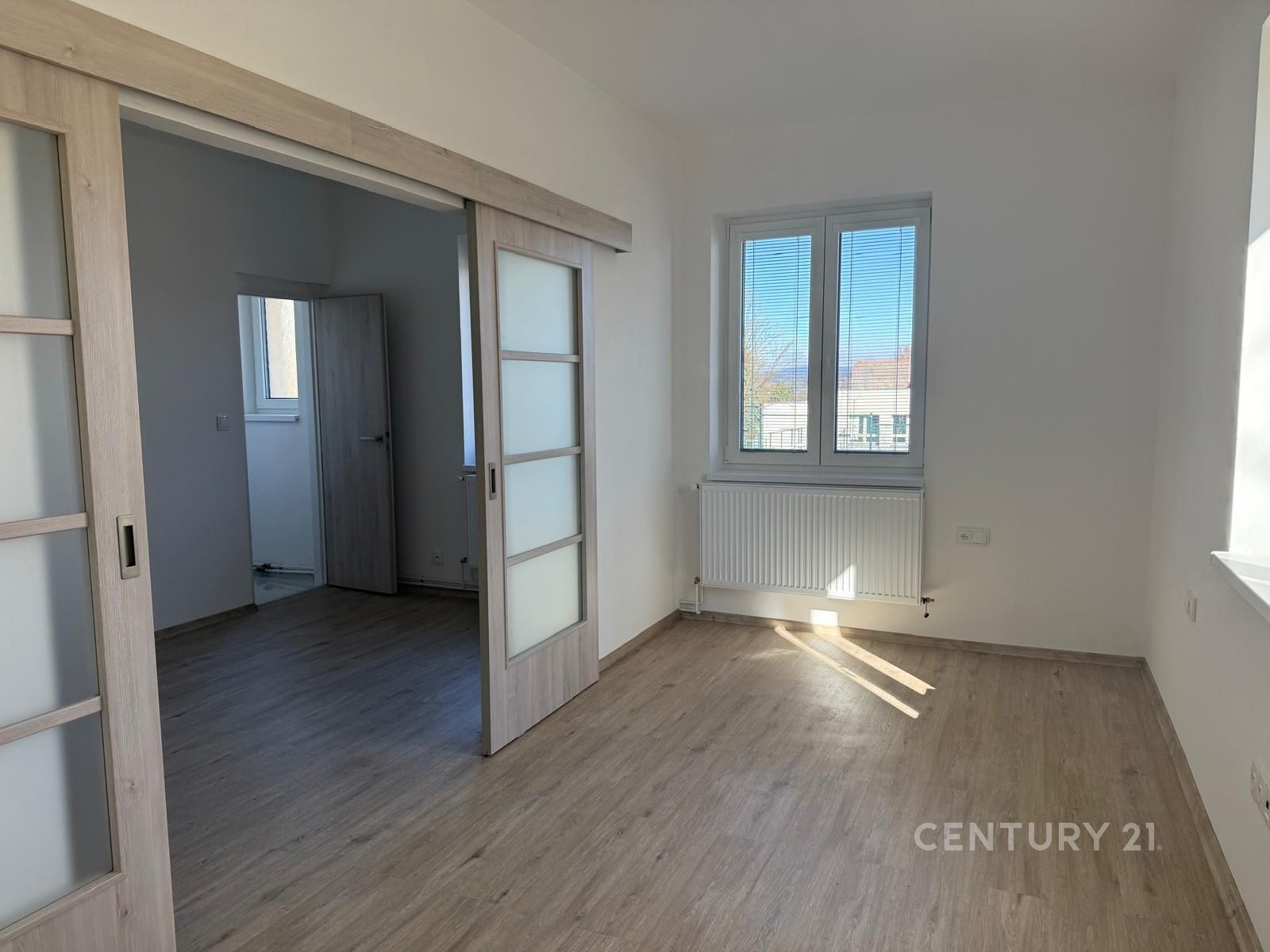 Světlý obývací pokoj s parketovou podlahou, velká okna poskytují přirozené světlo, moderní posuvné dveře oddělují místnosti.