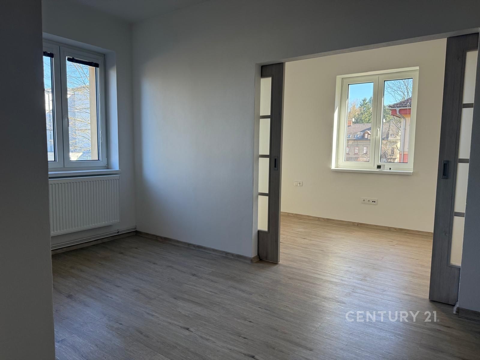 Světlá rekonstruovaná místnost s parketovou podlahou a velkými okny, které zajišťují dostatek denního světla.