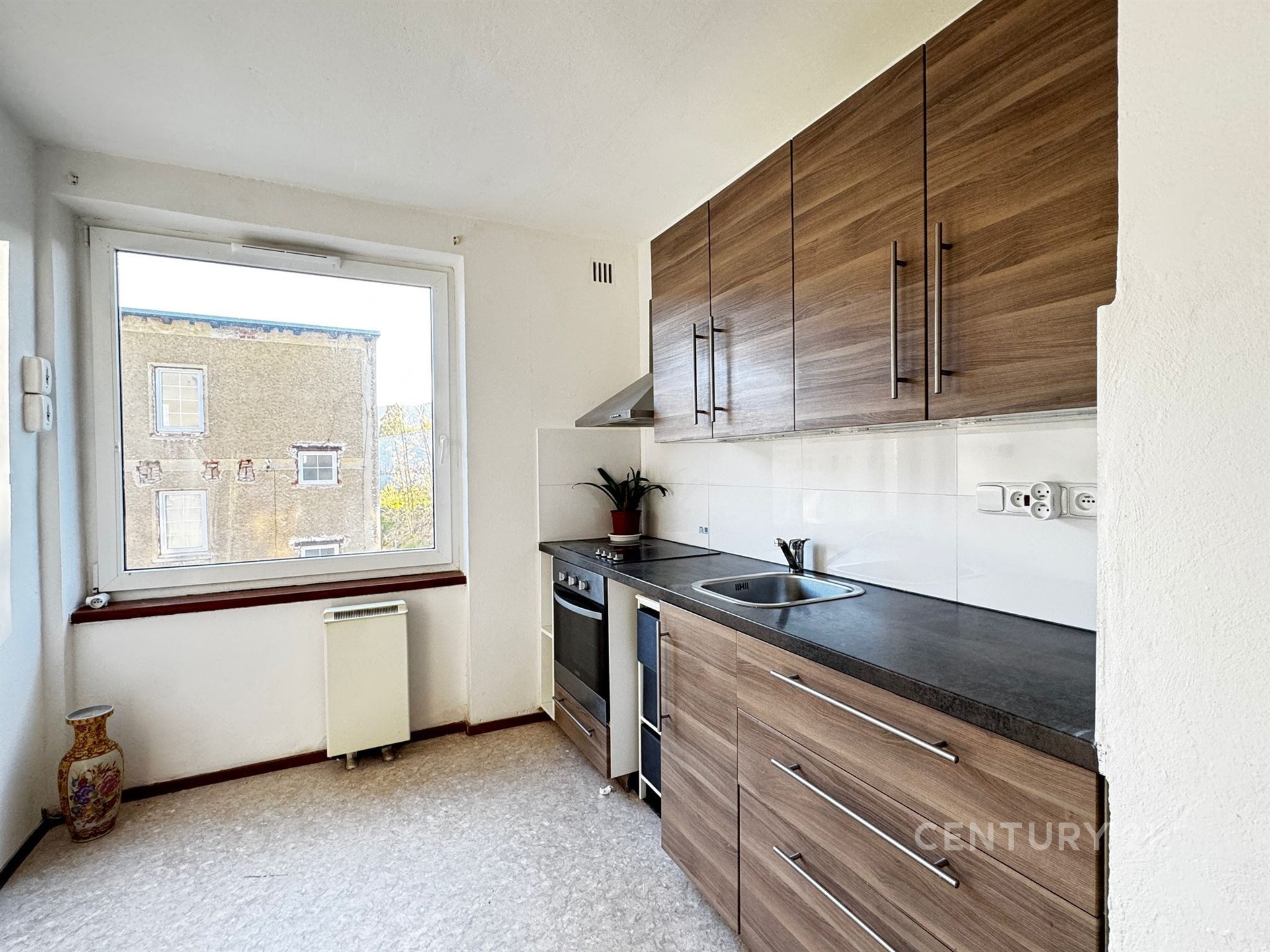 Kompaktní kuchyň s moderními dřevěnými skříňkami, velkým oknem a světlou dlažbou, ideální pro efektivní využití prostoru.