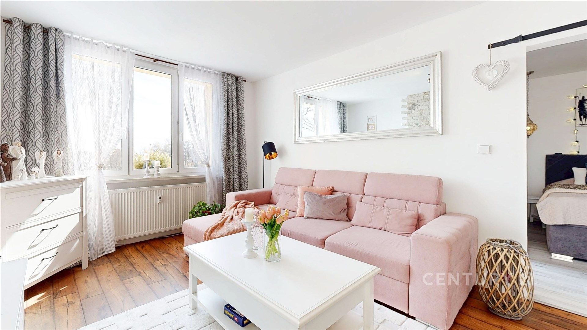 Světlý obývací pokoj s parketovou podlahou, velká okna a moderní design. Přirozené osvětlení a stylové dekorace vytvářejí útulnou atmosféru.