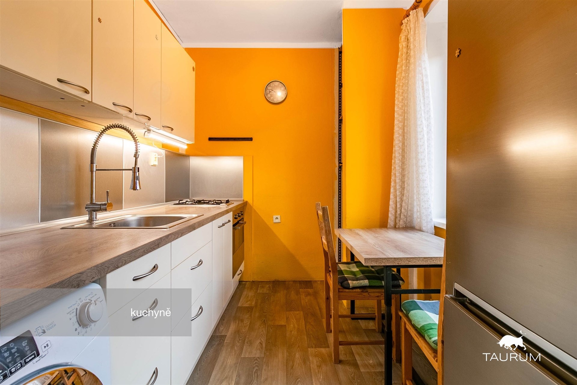 Kuchyně s dřevěnou podlahou, moderními spotřebiči a světlými skříňkami. Velké okno zajišťuje dostatek přirozeného světla.