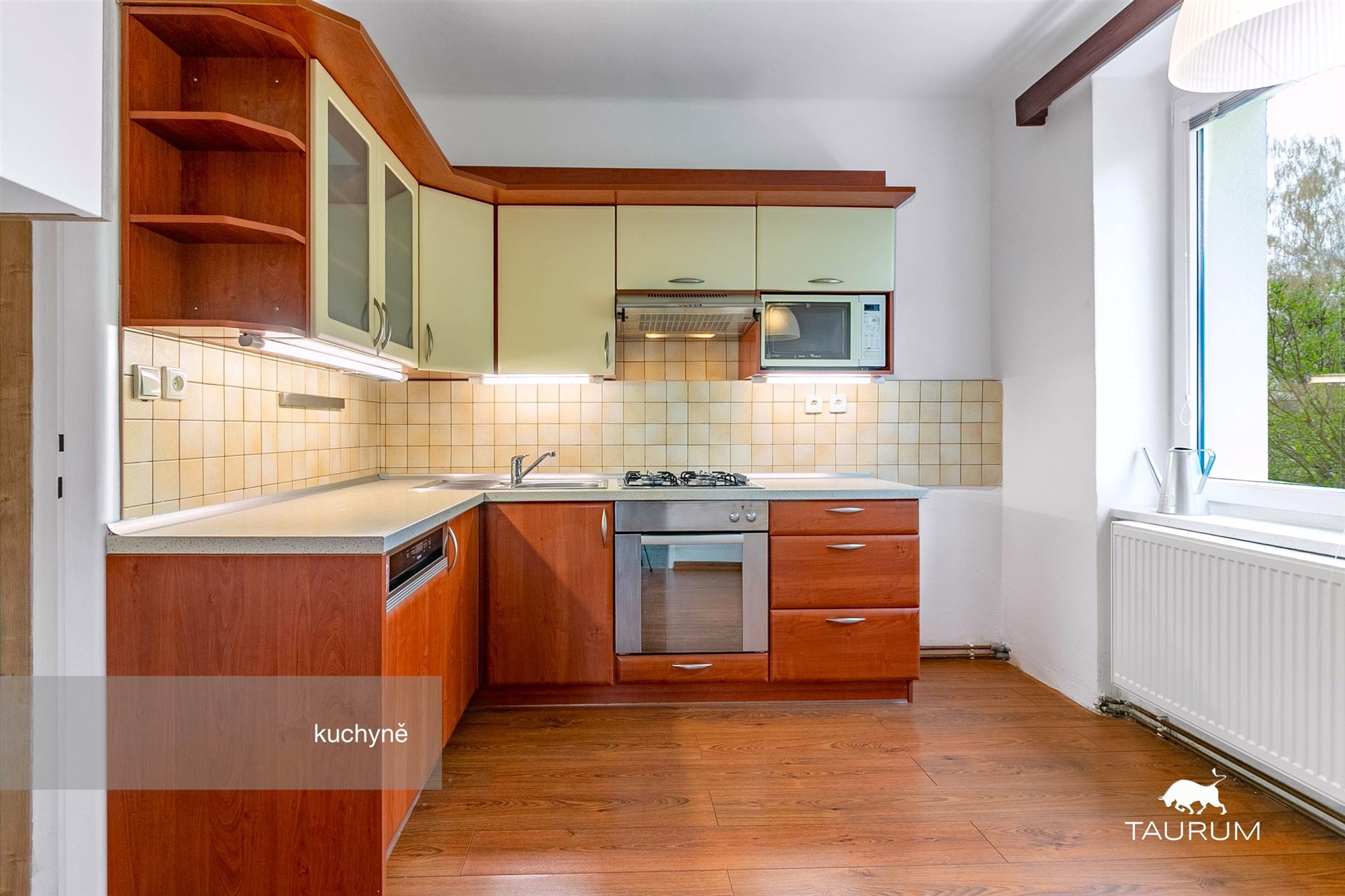 Moderní kuchyň s dřevěnou linkou, vestavěnými spotřebiči a parketovou podlahou, světlé dlaždice kolem pracovní desky, velké okno poskytuje dostatek světla