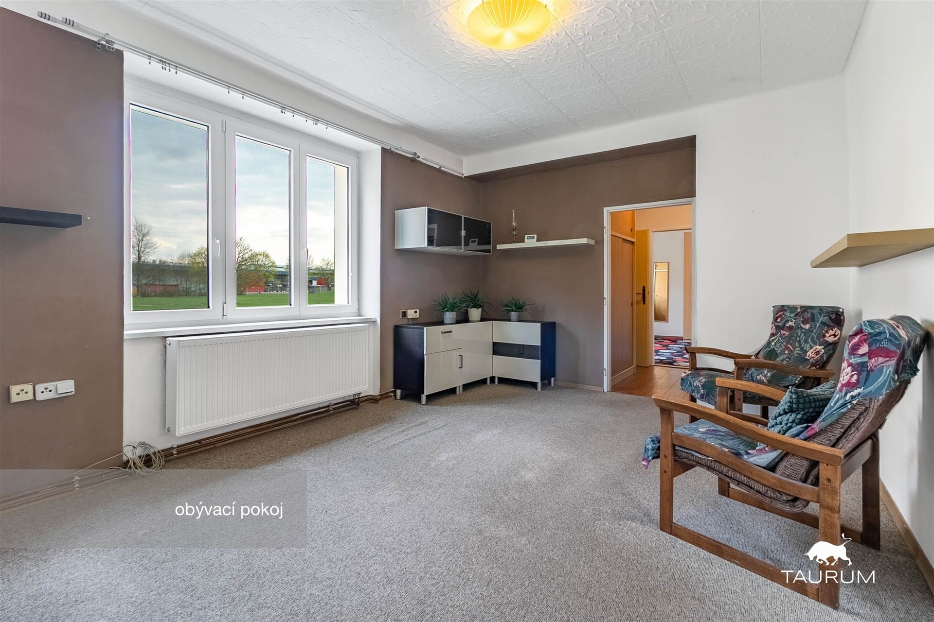 Obývací pokoj s velkými okny, kobercem a světlou stěnou, nabízí dostatek přirozeného světla a komfortní prostor pro relaxaci.