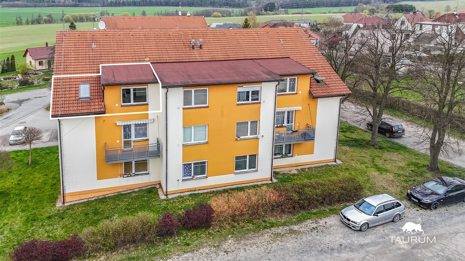 Malý bytový dům s oranžovou fasádou, renovovaná střecha z pálených tašek, klidná lokalita uprostřed zeleně, parkování před domem.