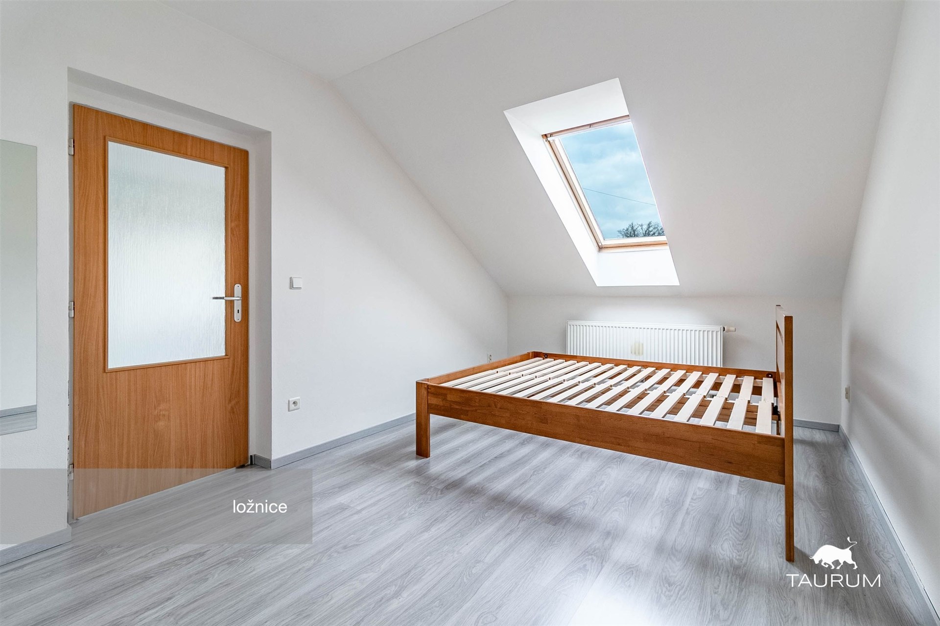 Prostorná ložnice s laminátovou podlahou, velké střešní okno, moderní interiér s dřevěným rámem postele, světlý a vzdušný prostor.